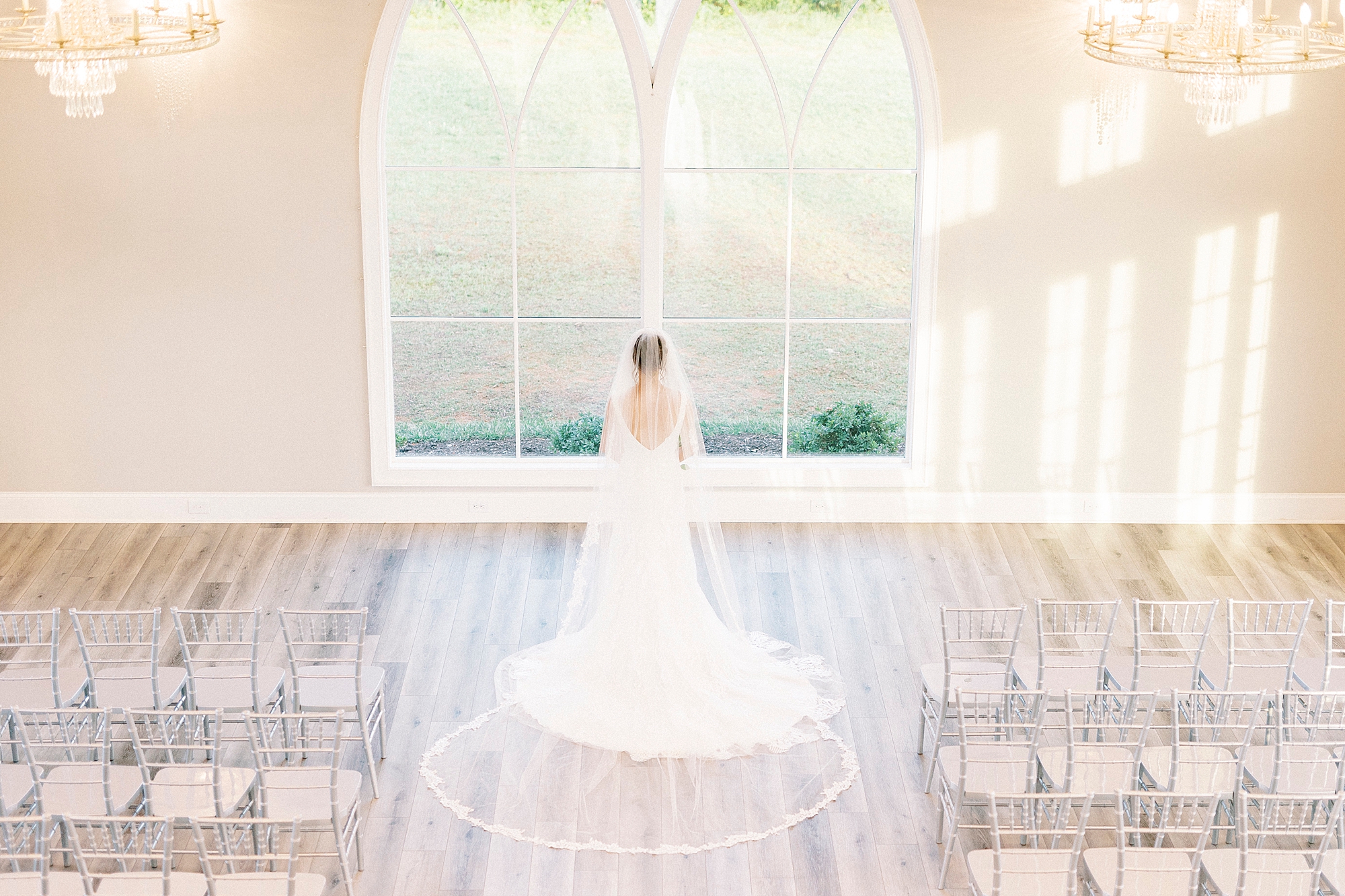 bride stands in chapel at Chickadee Hill Farms facing window with veil behind her 
