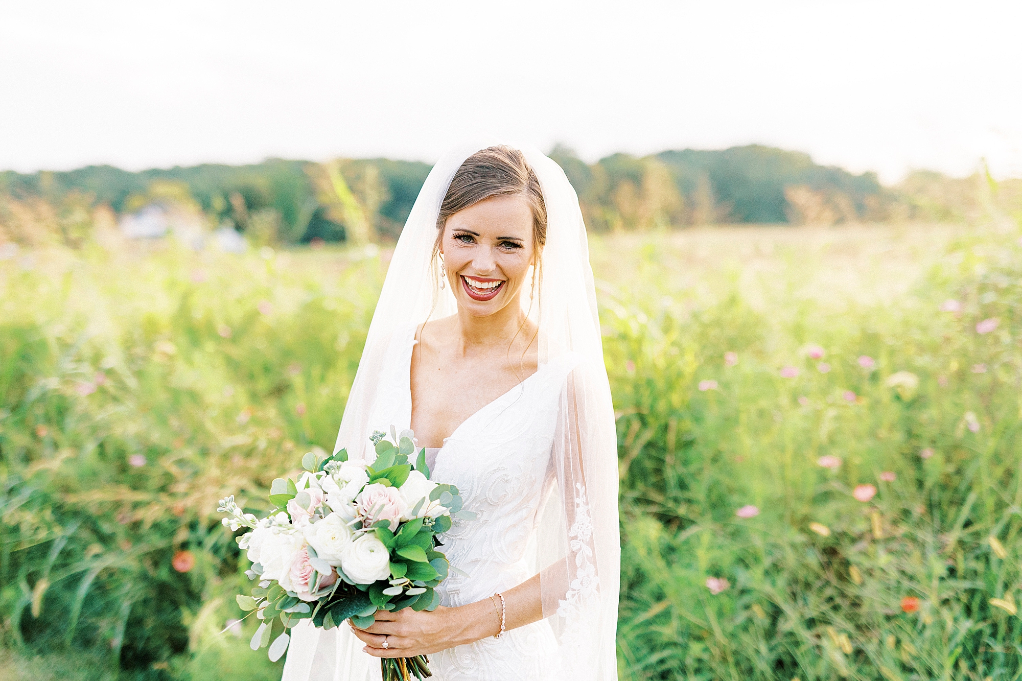 bride smiles in field holding bouquet with veil around her at Chickadee Hill Farms
