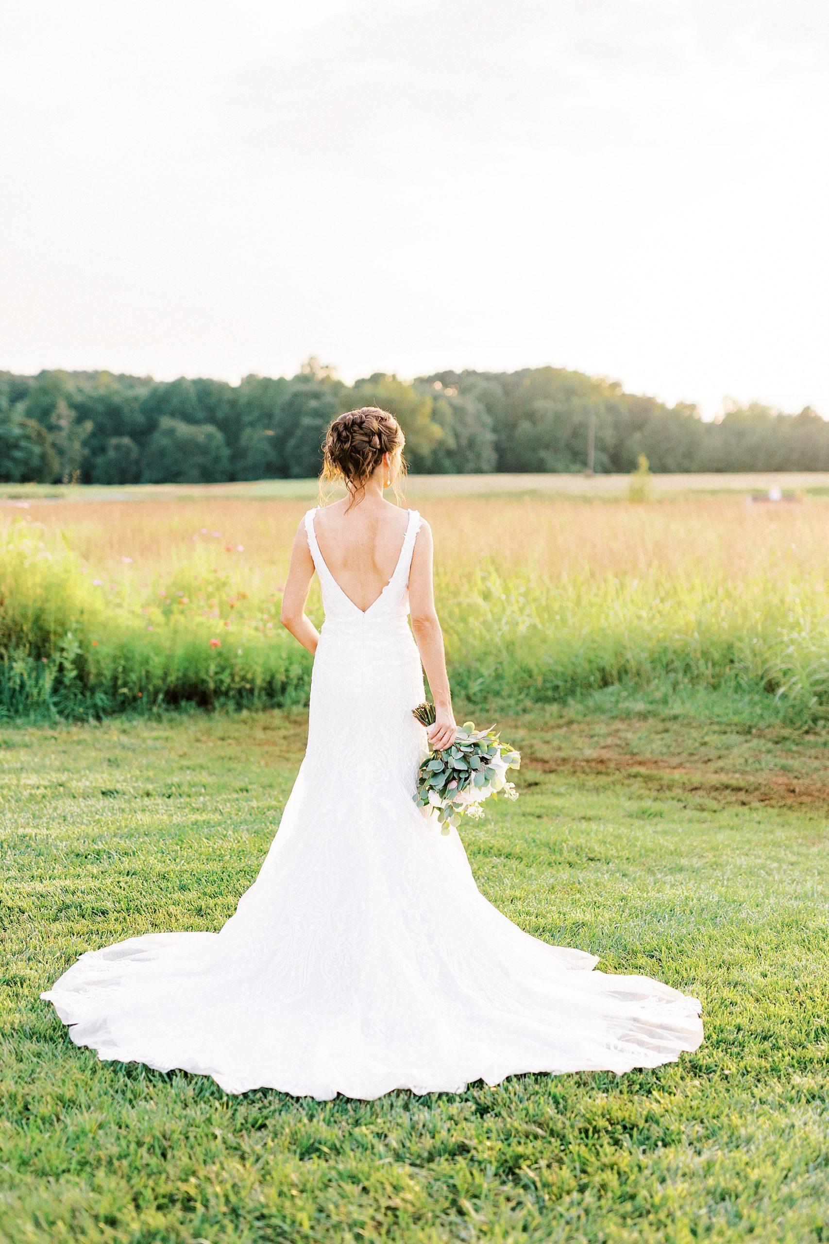 bride stands looking at field at Chickadee Hill Farms showing off back of gown