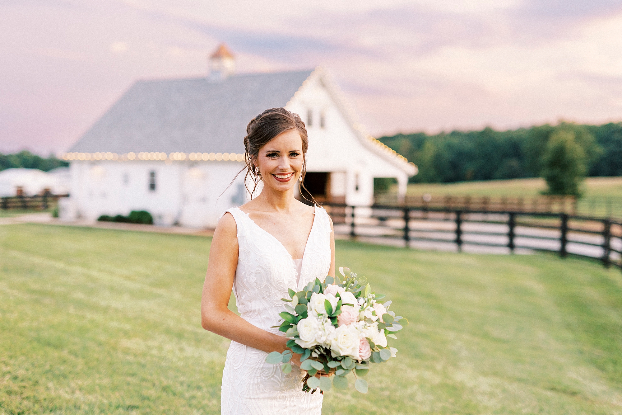 bride poses by white barn at Chickadee Hill Farms at sunset