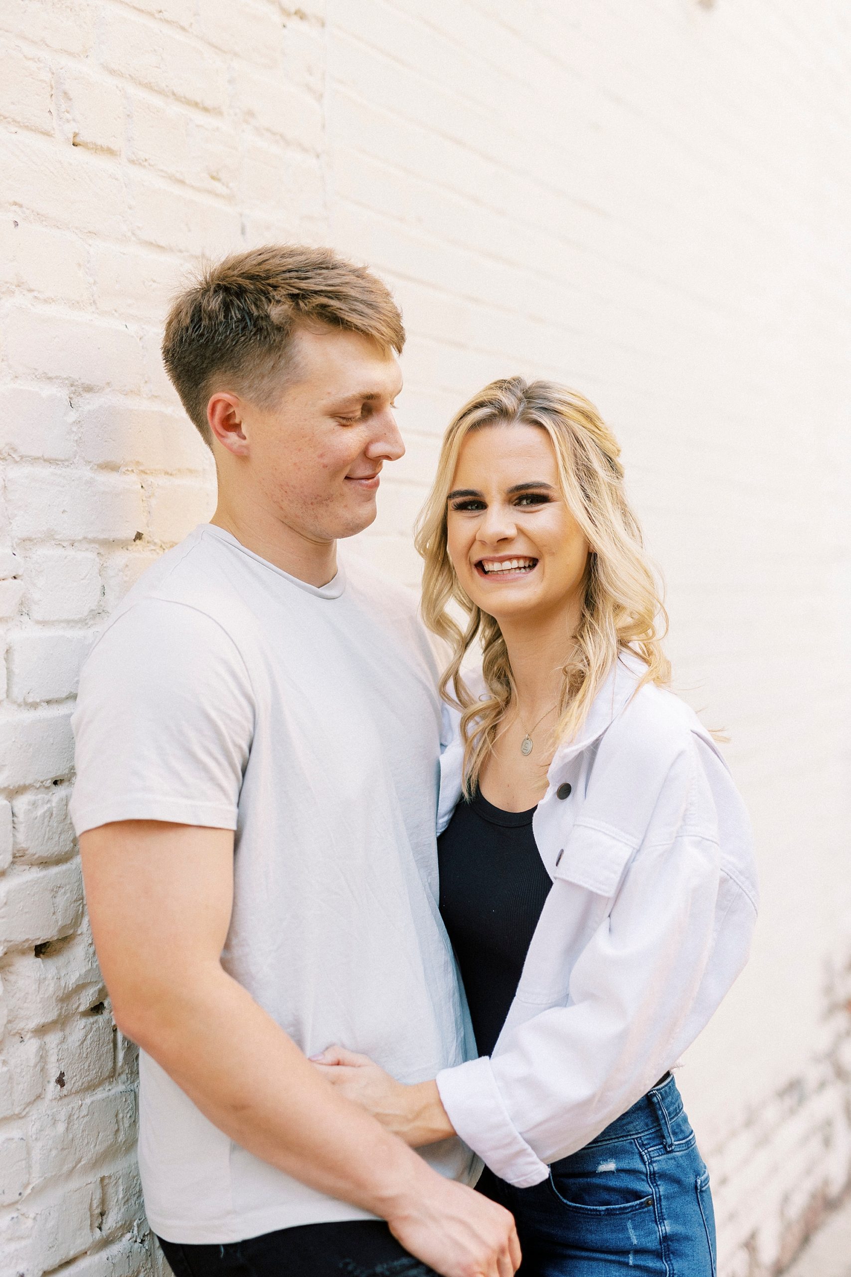 man looks down at fiancee leaning against white washed walls during Downtown Concord engagement portraits