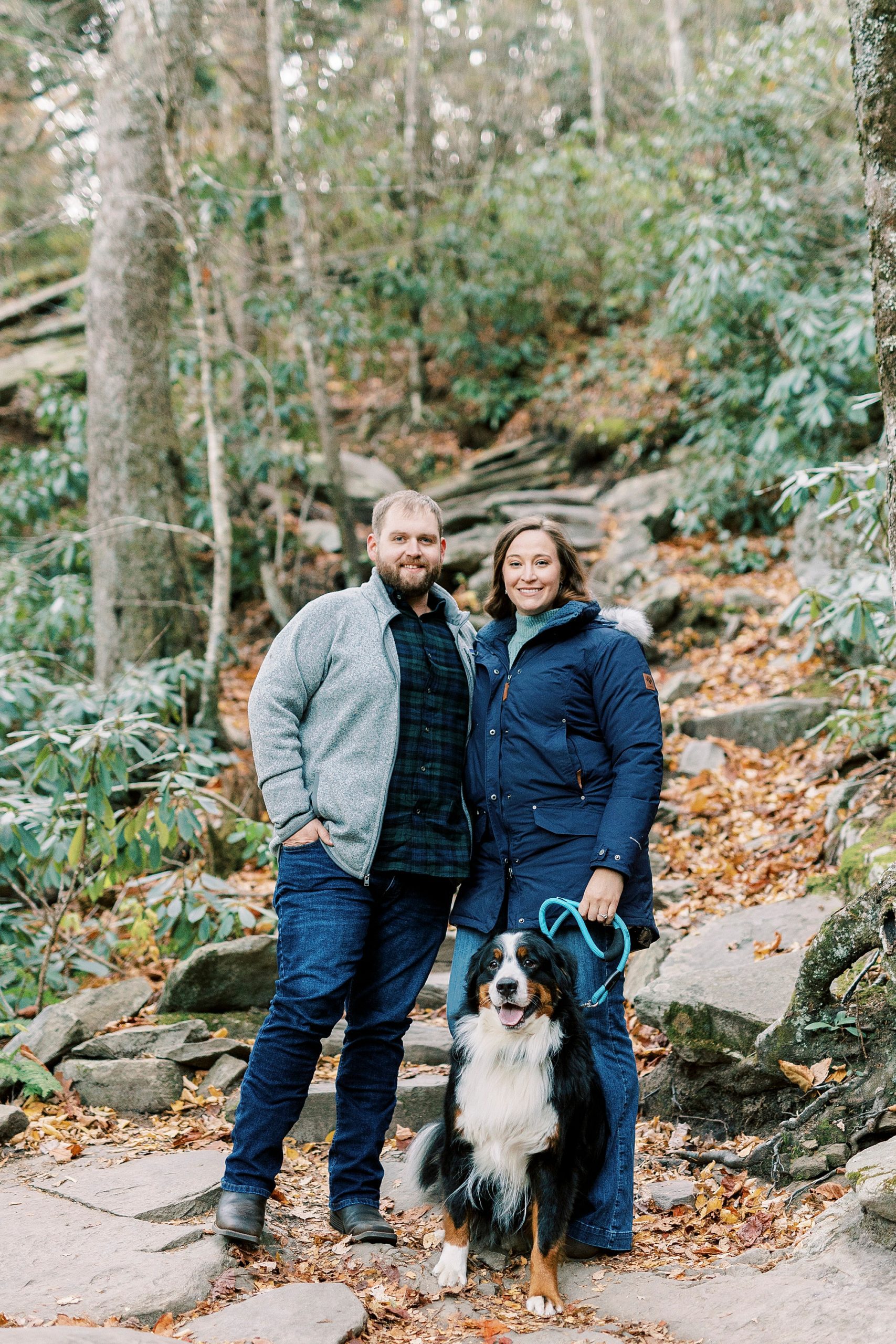 couple stands with dog during portraits at base of Rough Ridge