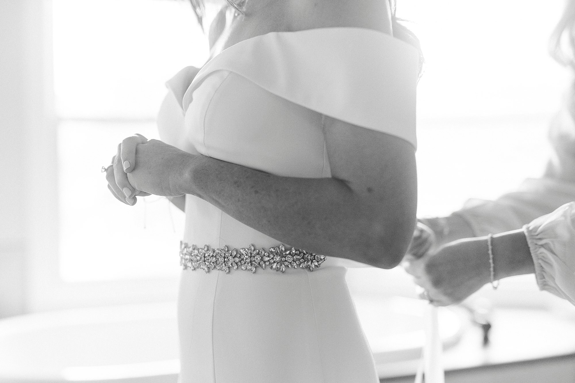 bride in off-the-shoulder gown gets ribbon tied around waist 