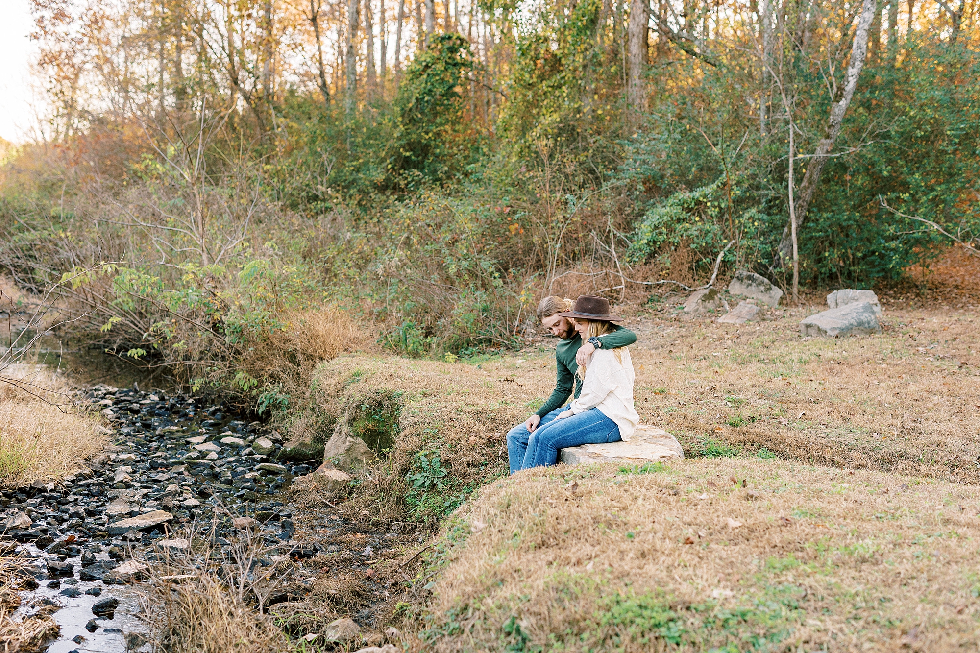 bride and groom sit together by creek during chic fall engagement session