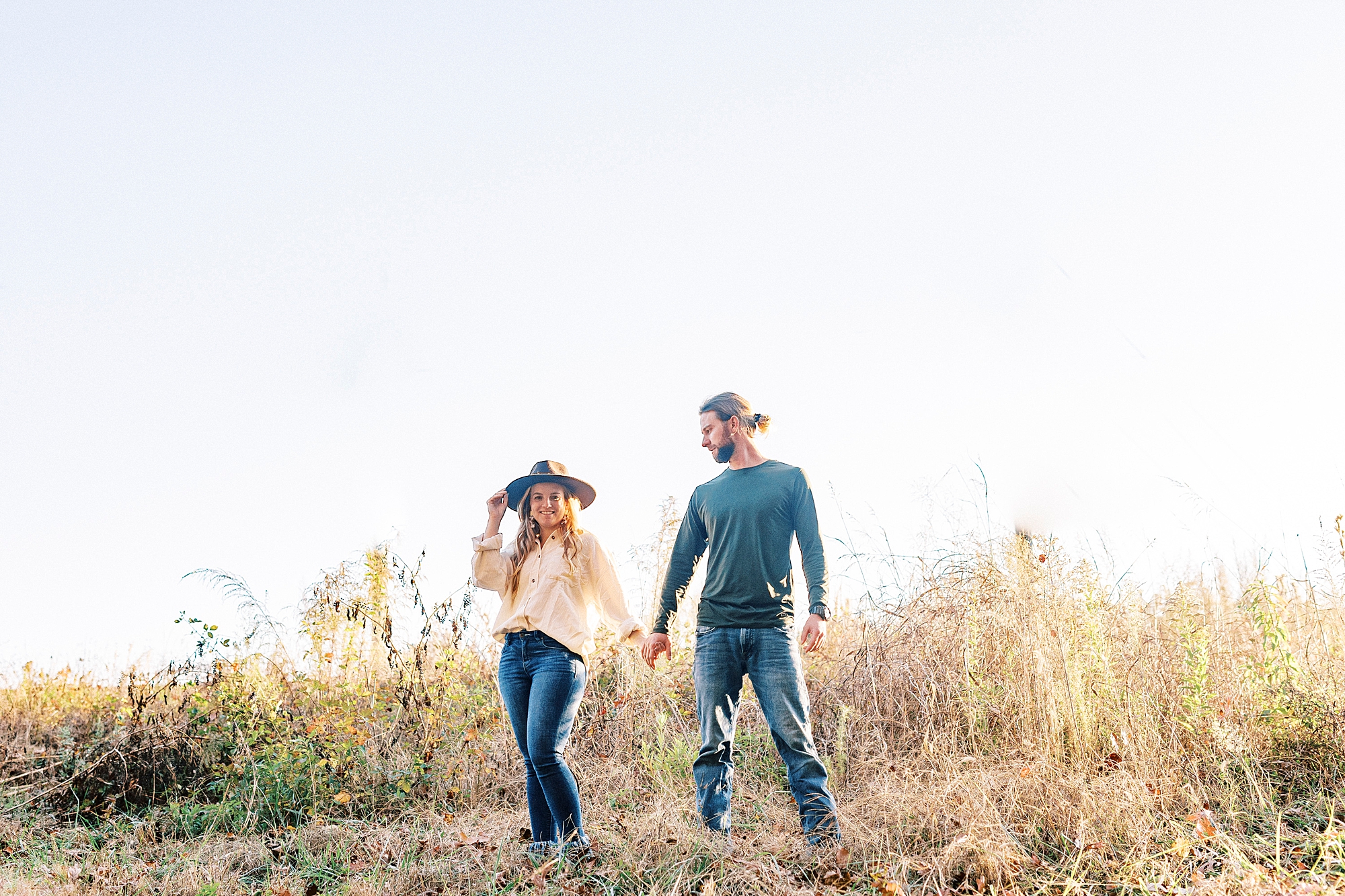 engaged couple holds hands walking through field during chic fall engagement session
