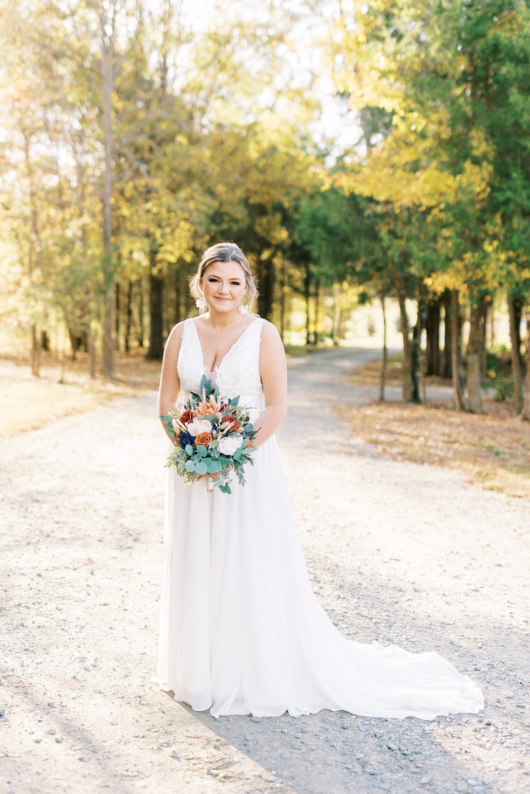 fall bride stands on driveway with bouquet of orange and peach flowers 