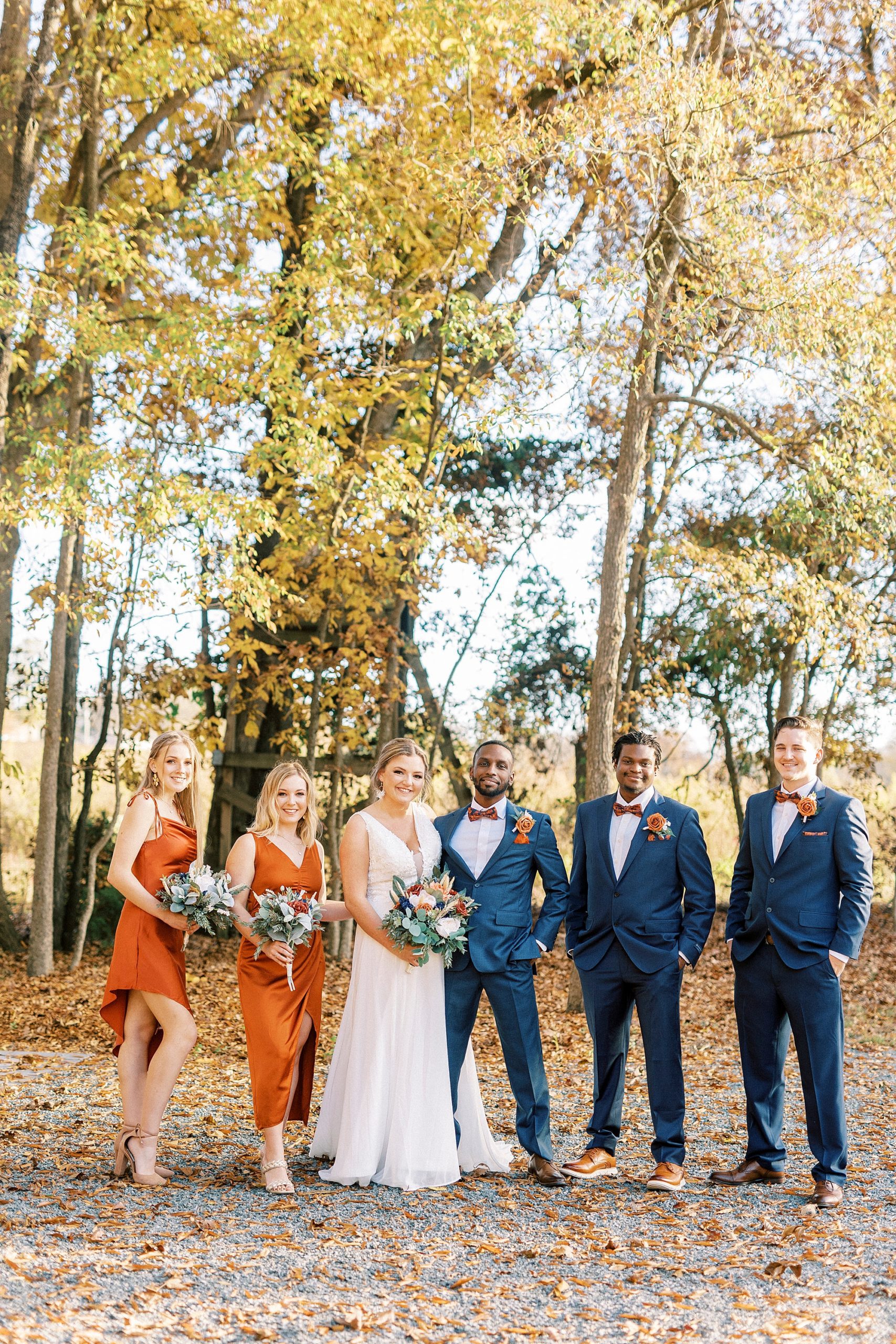 newlyweds stand together with bridesmaids in burnt orange and groomsmen in navy suits 