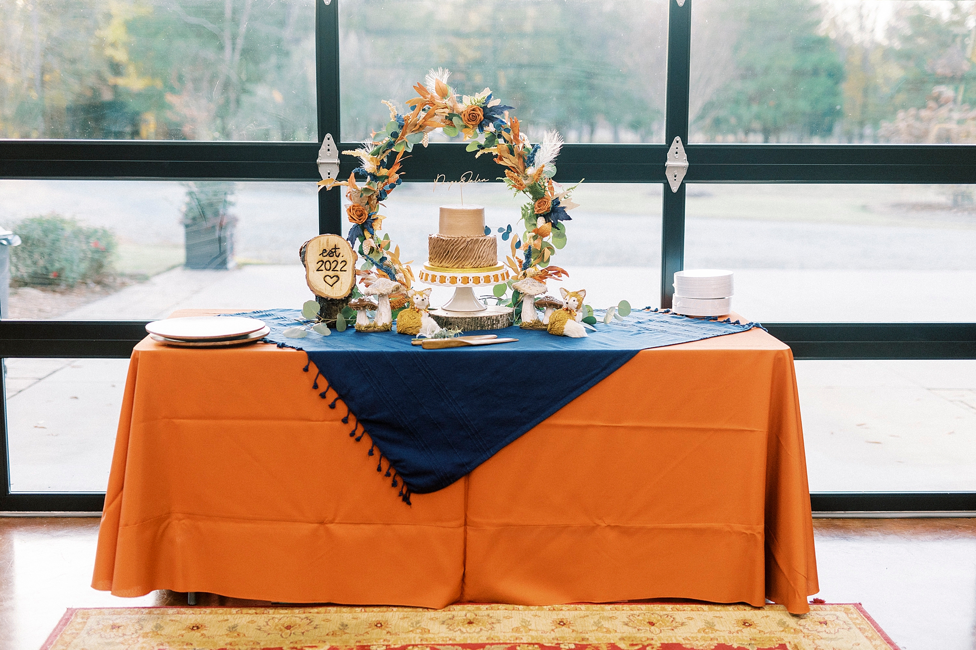 wedding cake sits on table with orange table cloth and floral arch 