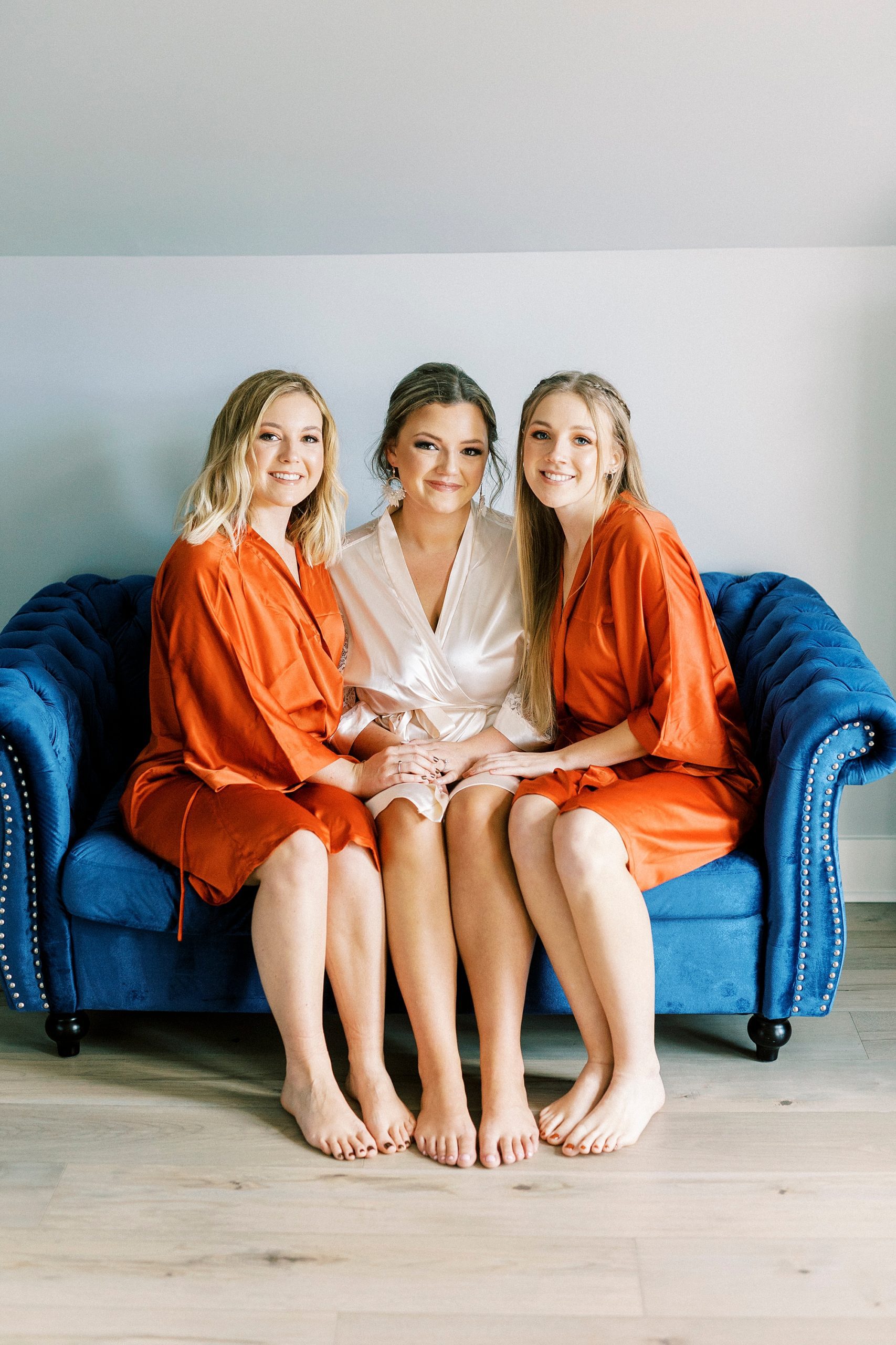 bride sits on blue couch with bridesmaids in orange robes 