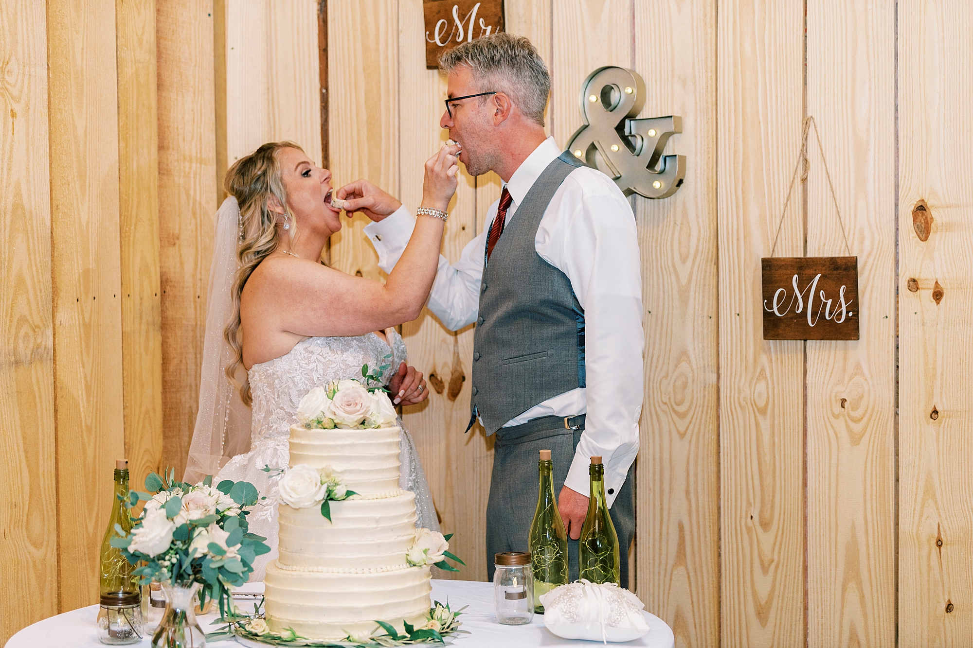 bride and groom feed each other cake by wooden wall of barn 