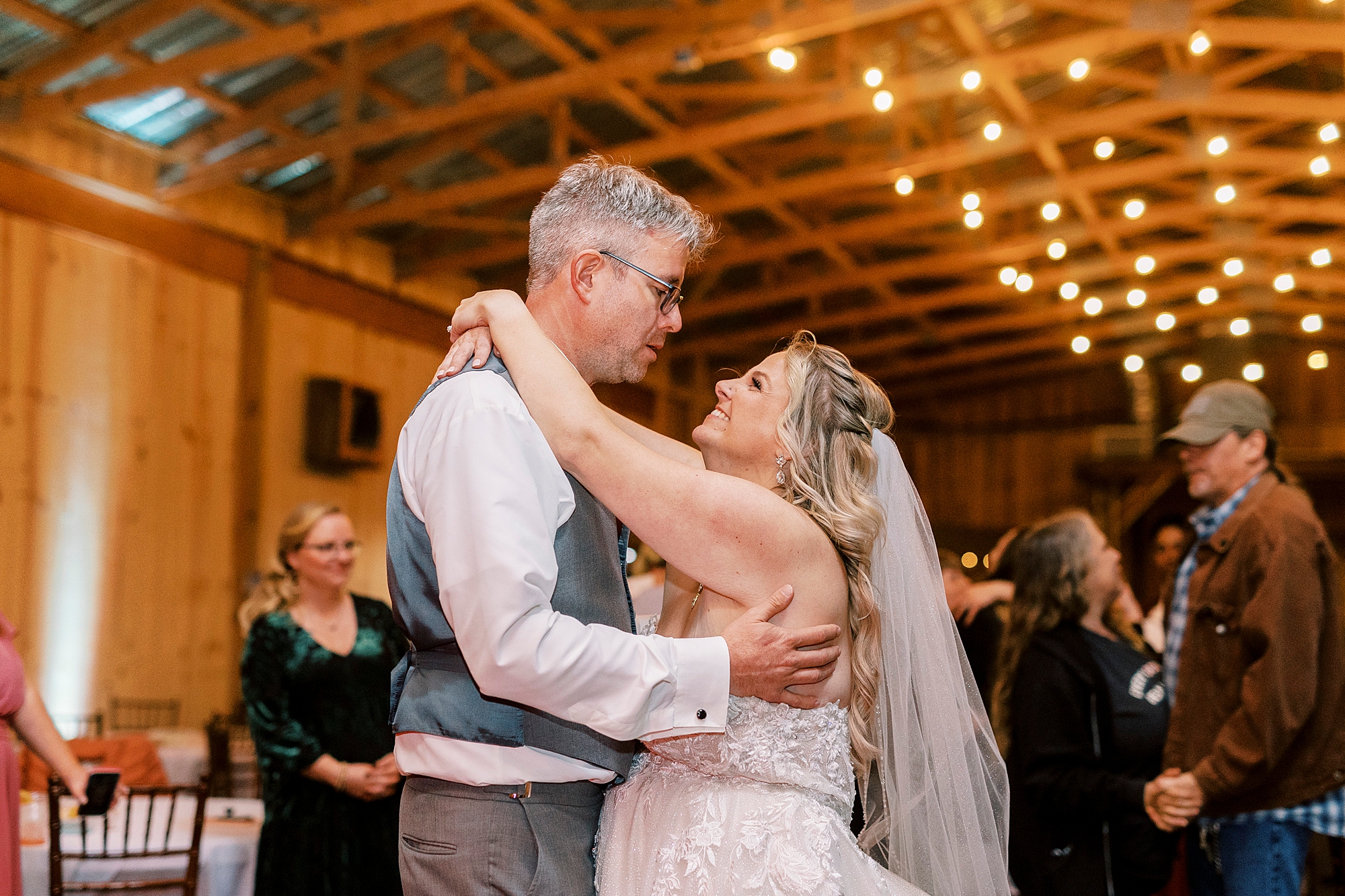 bride hugs groom around shoulders looking at him during reception in barn at the Gin at Aw Shucks Farm