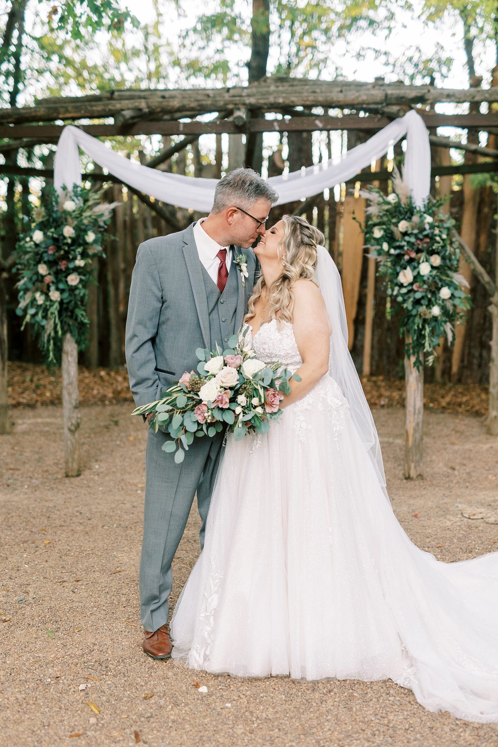bride and groom smile touching noses under wooden arbor at the Gin at Aw Shucks Farm