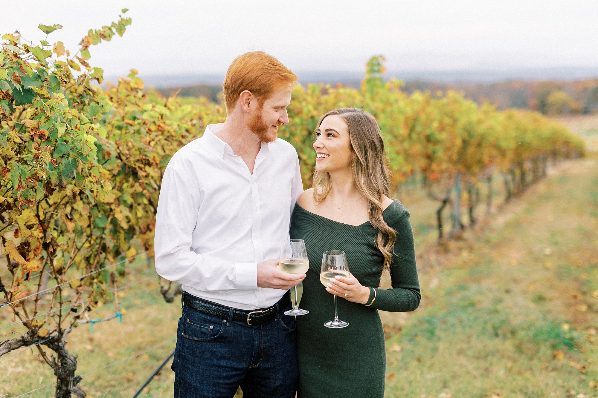 engaged couple holds glass of wine between vines