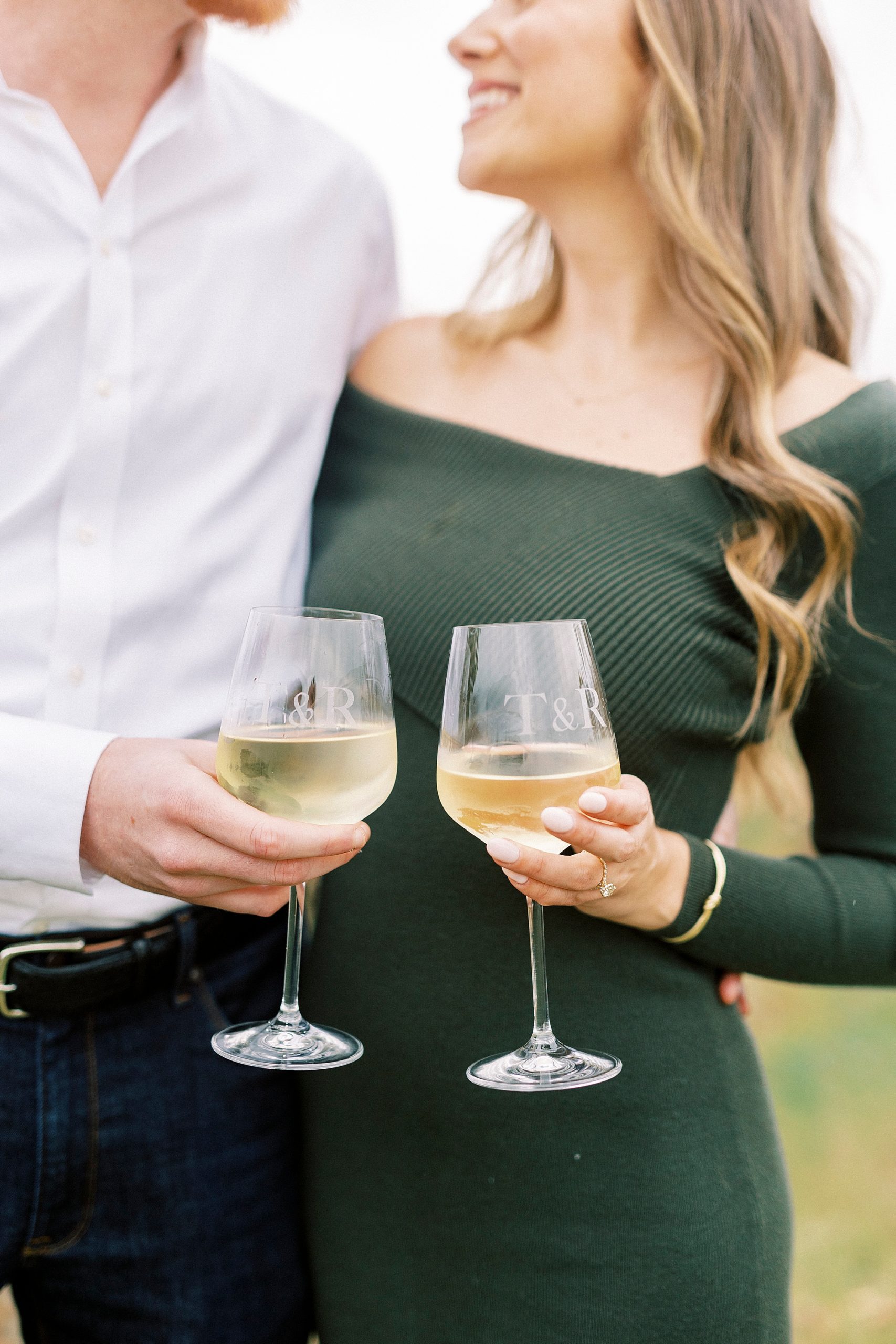 woman in chic green dress holds glass of white wine with fiancee hugging her 