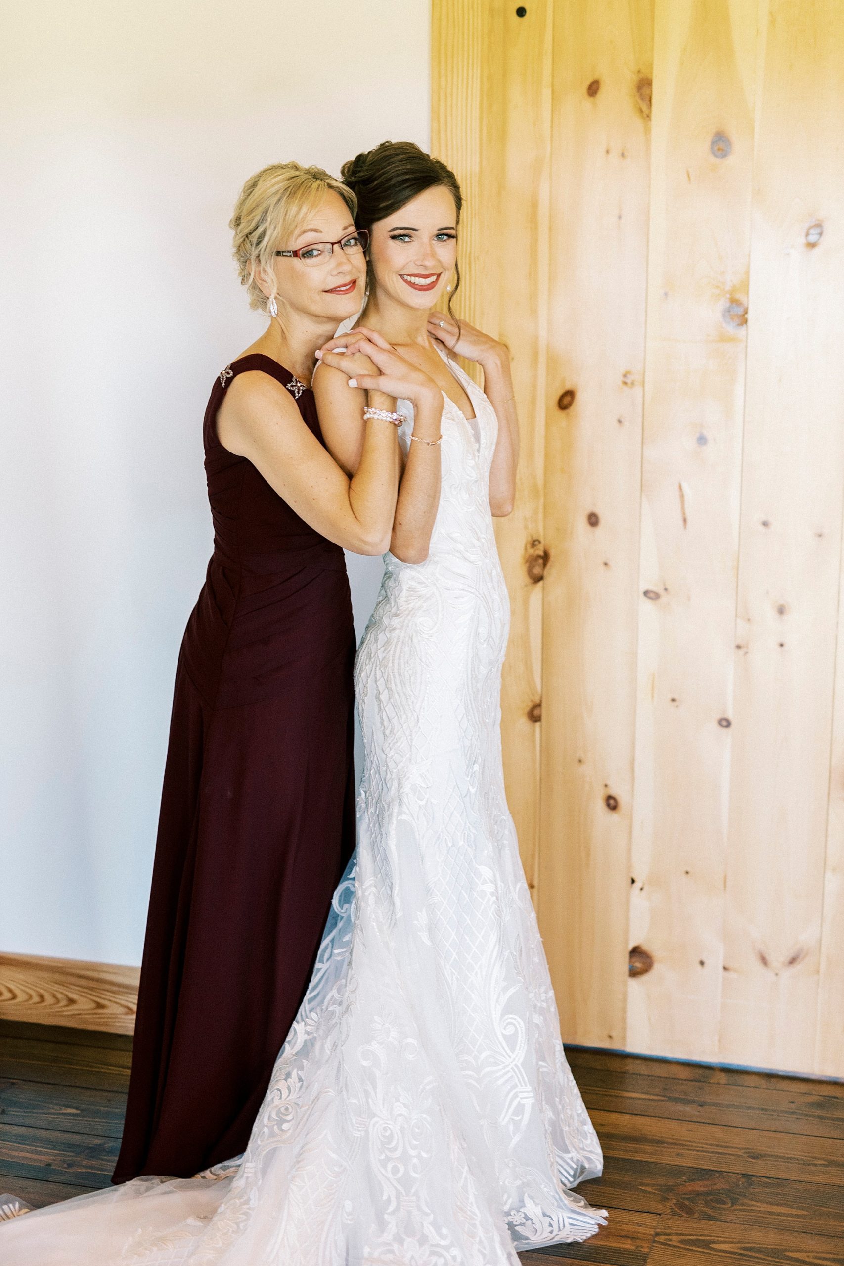 mother in burgundy gown hugs bride during prep at Chickadee Hill Farms