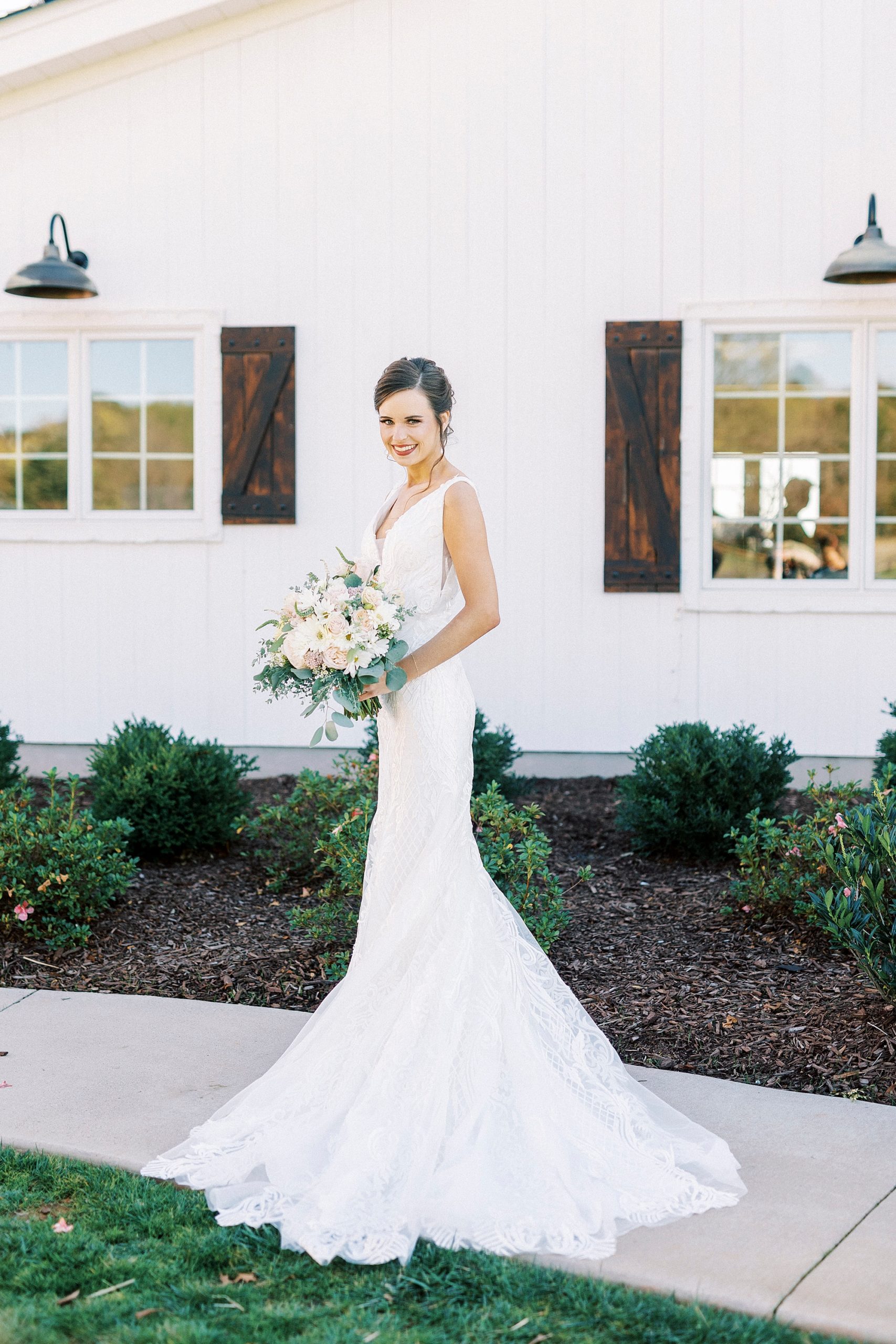 bride poses on sidewalk by white barn at Chickadee Hill Farms