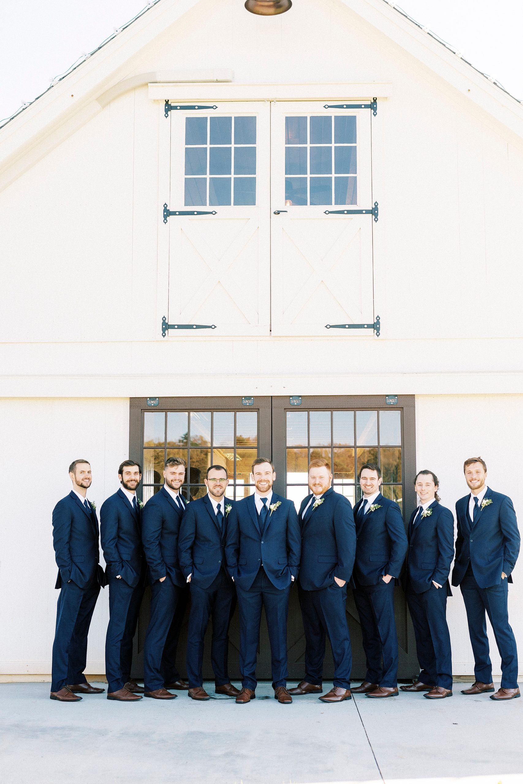 groom and groomsmen in navy suits pose by wooden doors at Chickadee Hill Farms