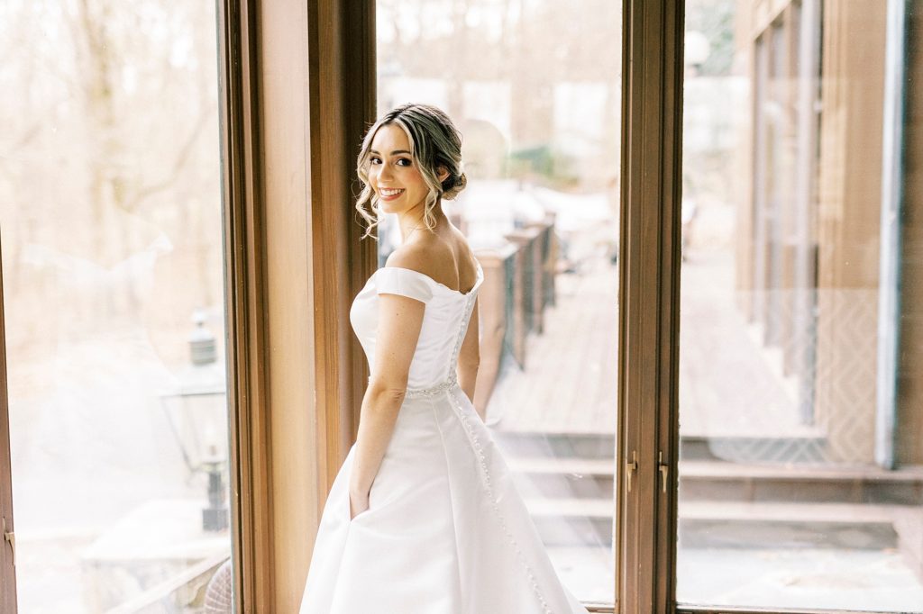 bride stands with hands in pocket of wedding gown smiling over shoulder 