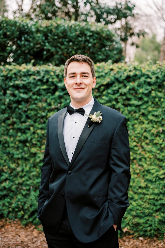 groom poses in black suit by green hedge 