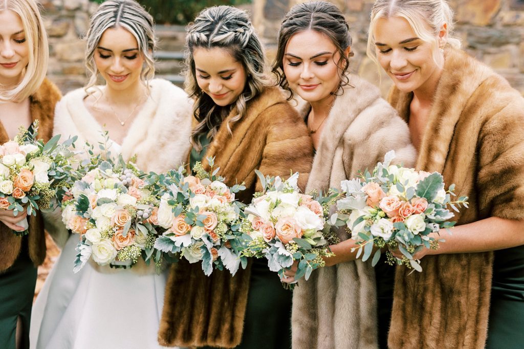 bride poses with bridesmaids in emerald green gowns with brown furs looking at bouquets 