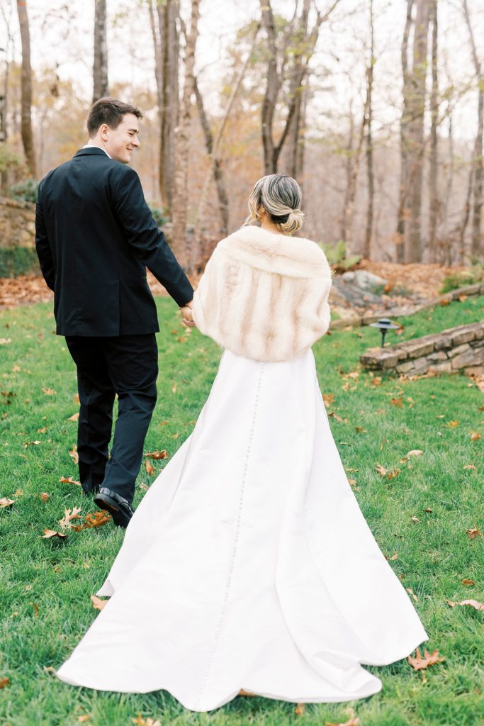 bride holds groom's hand walking on lawn in fur 