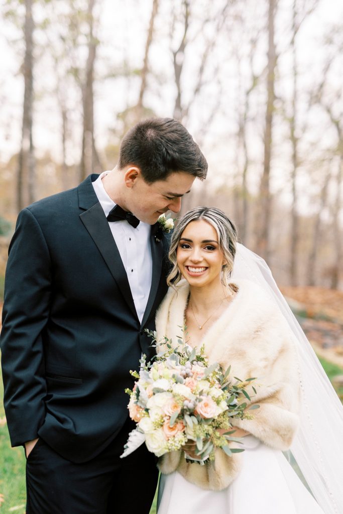 groom in black suit smiles down at bride in light brown fur 
