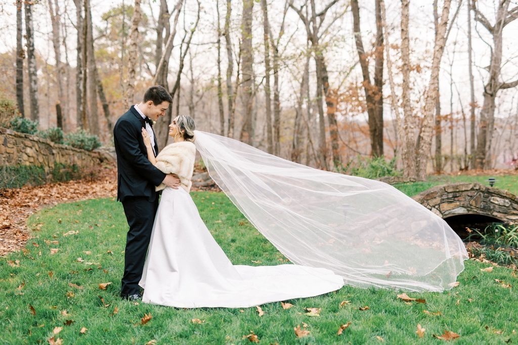 bride and groom smile together with her veil floating behind them on lawn 
