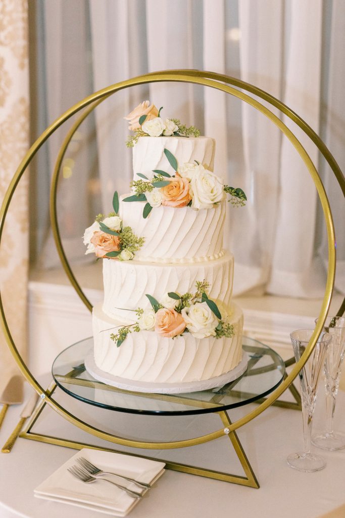 tiered wedding cake sits in gold display with peach and white flowers 