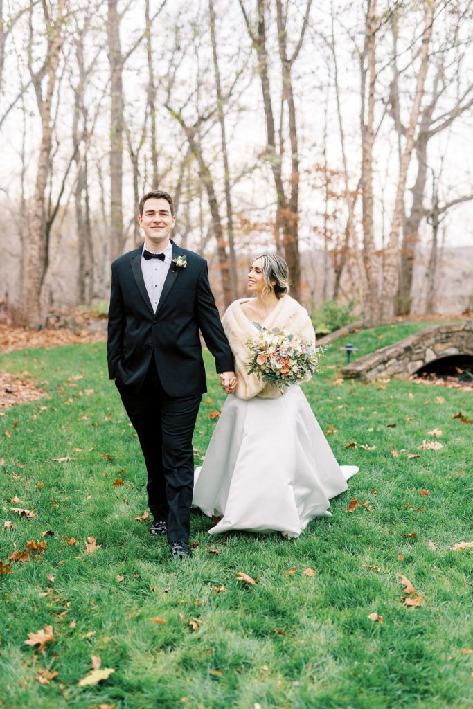 bride and groom hold hands walking across lawn with fallen leaves