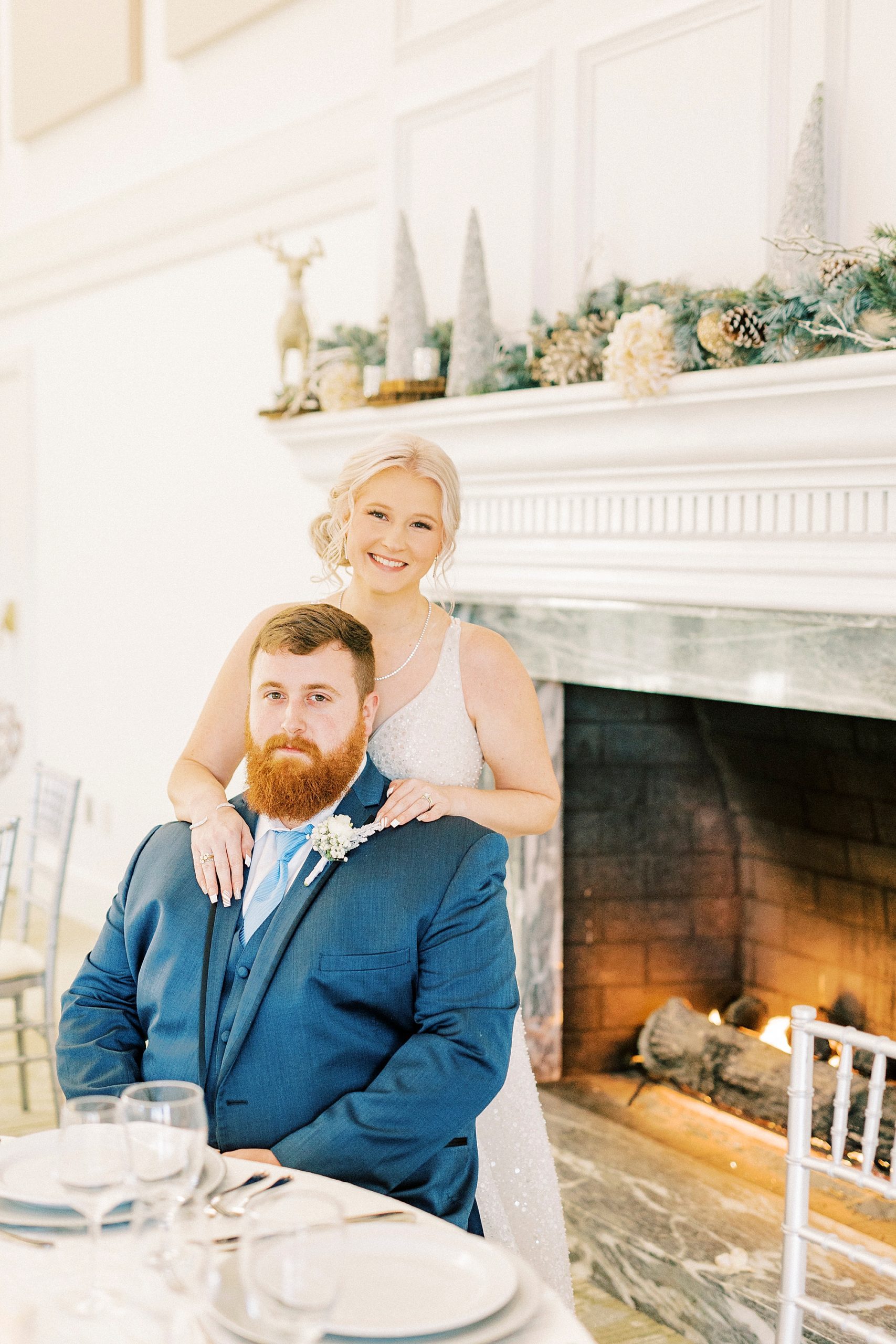 bride stands behind groom in front of fireplace at River Run Country Club