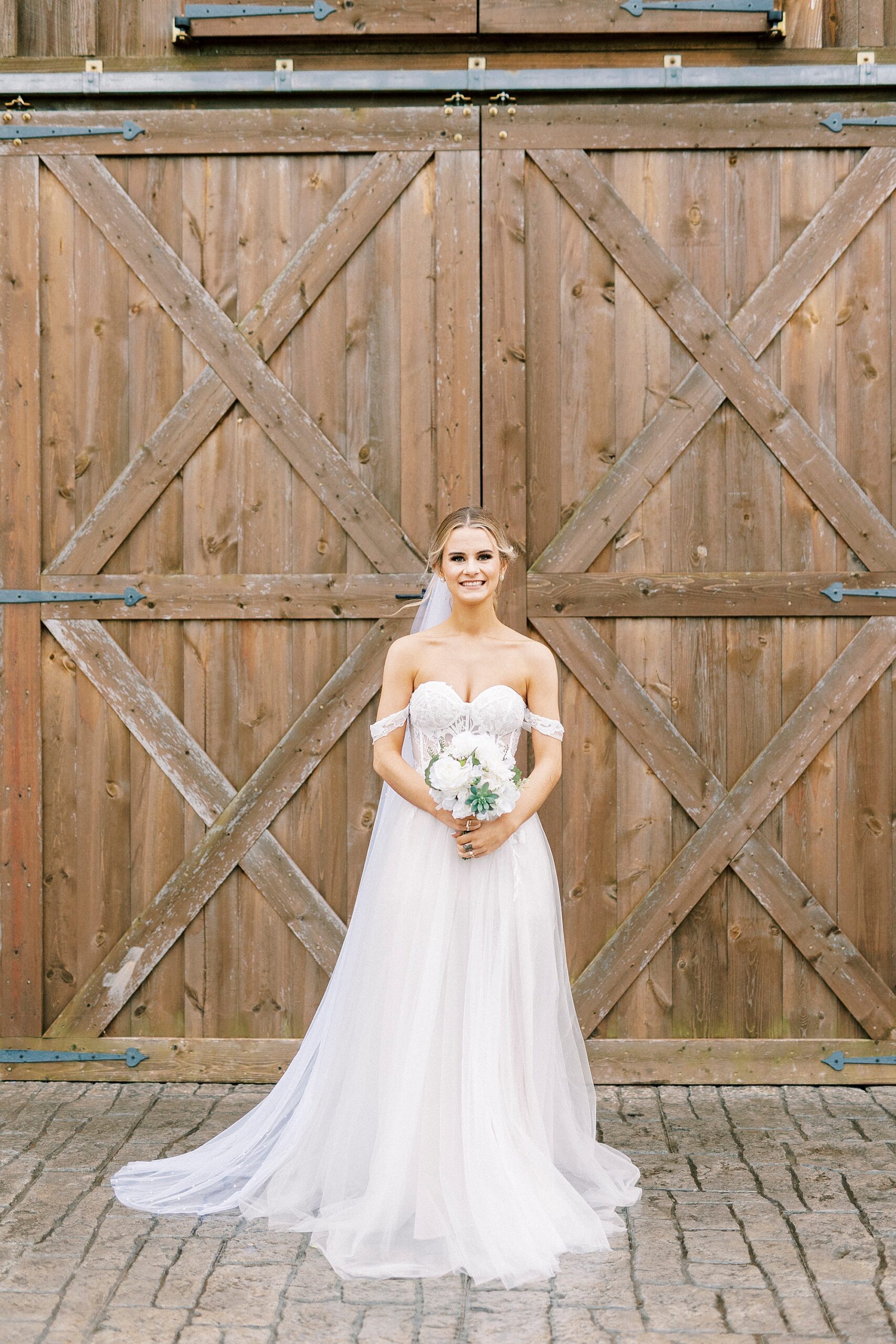 bride poses by wooden barn doors at The Farmstead holding white bouquet with succulents 