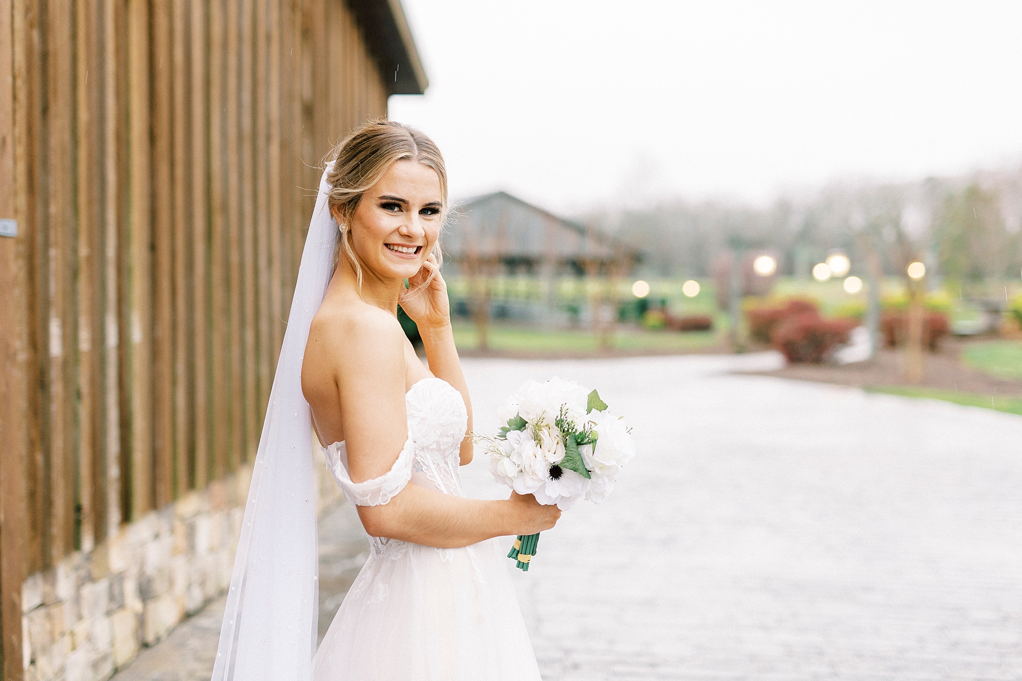 bride smiles looking over shoulder by wooden barn during bridal portraits at The Farmstead