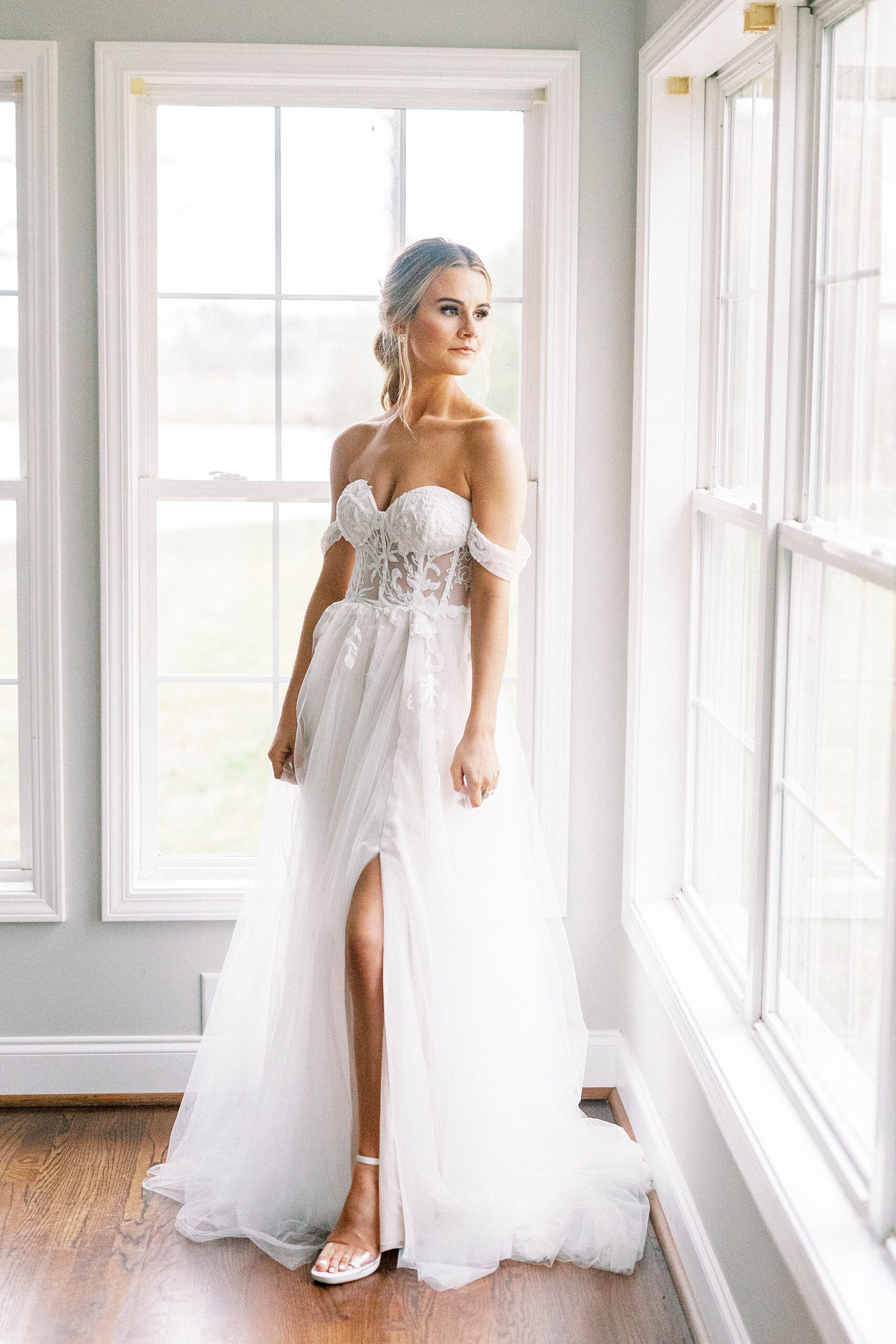bride in off-the-shoulder wedding gown with corset bodice looks over shoulder out window at The Farmstead