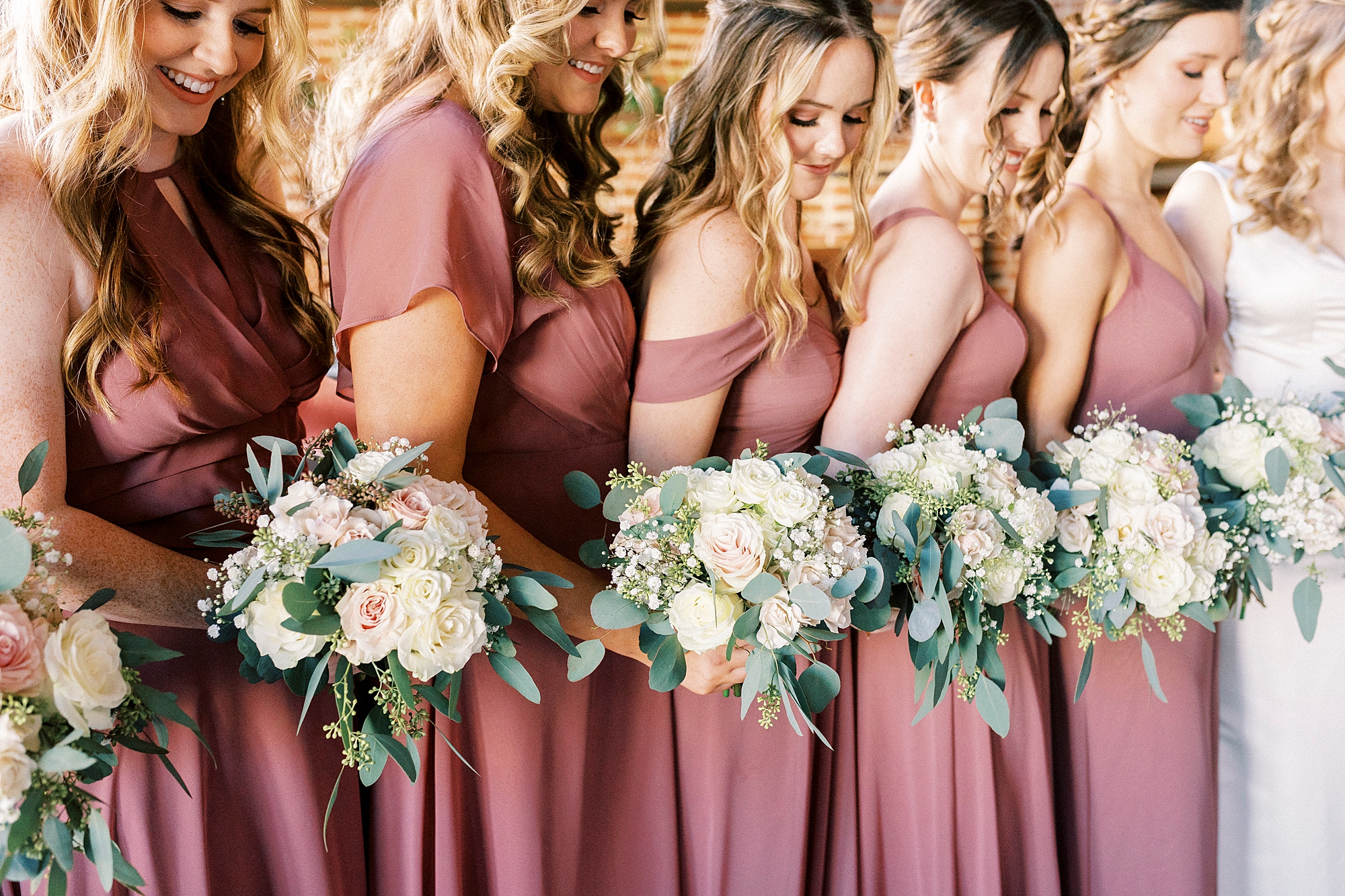 bridesmaids in pink gowns look down at bouquet of white flowers 
