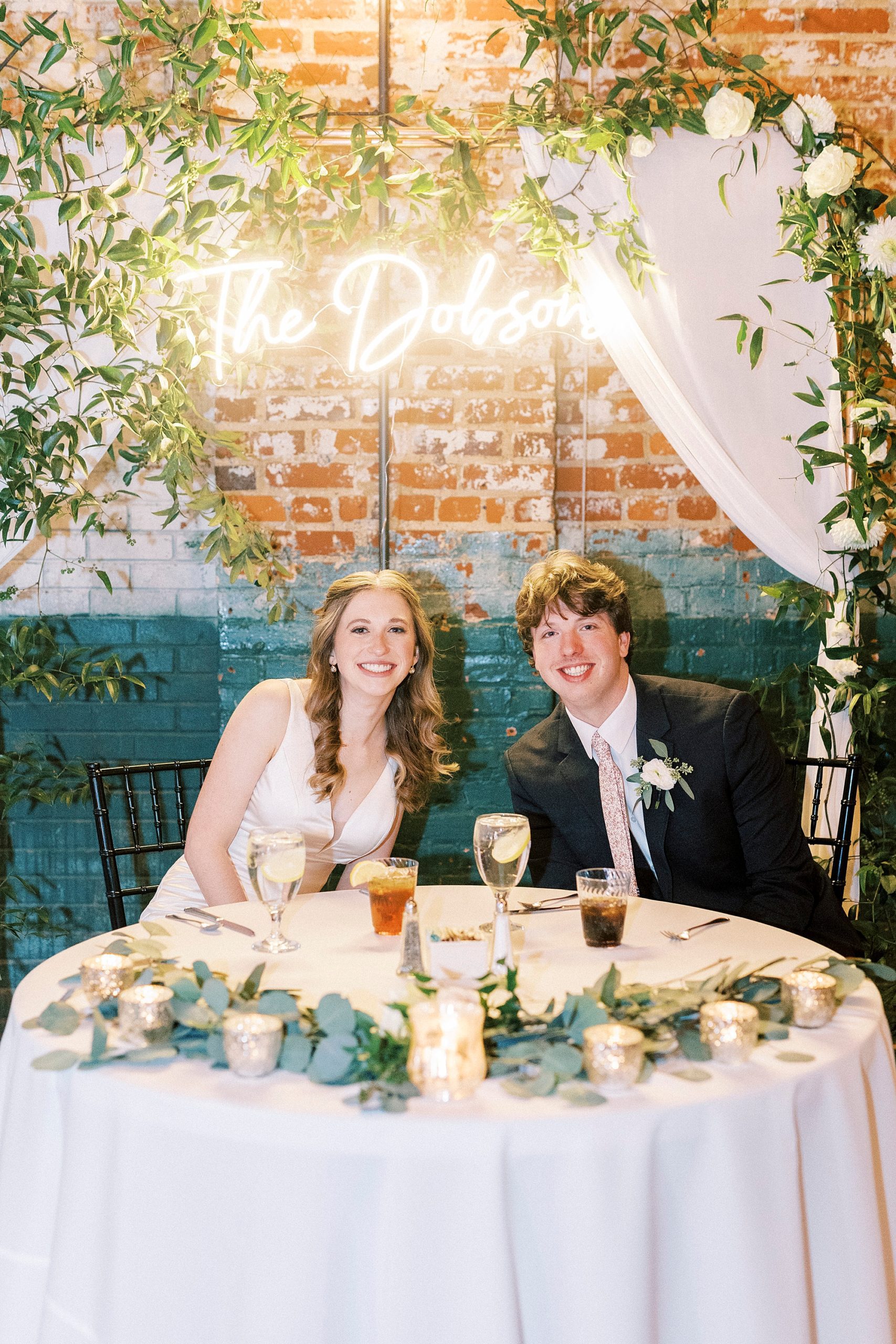 bride and groom sit at sweetheart table under custom neon sign 