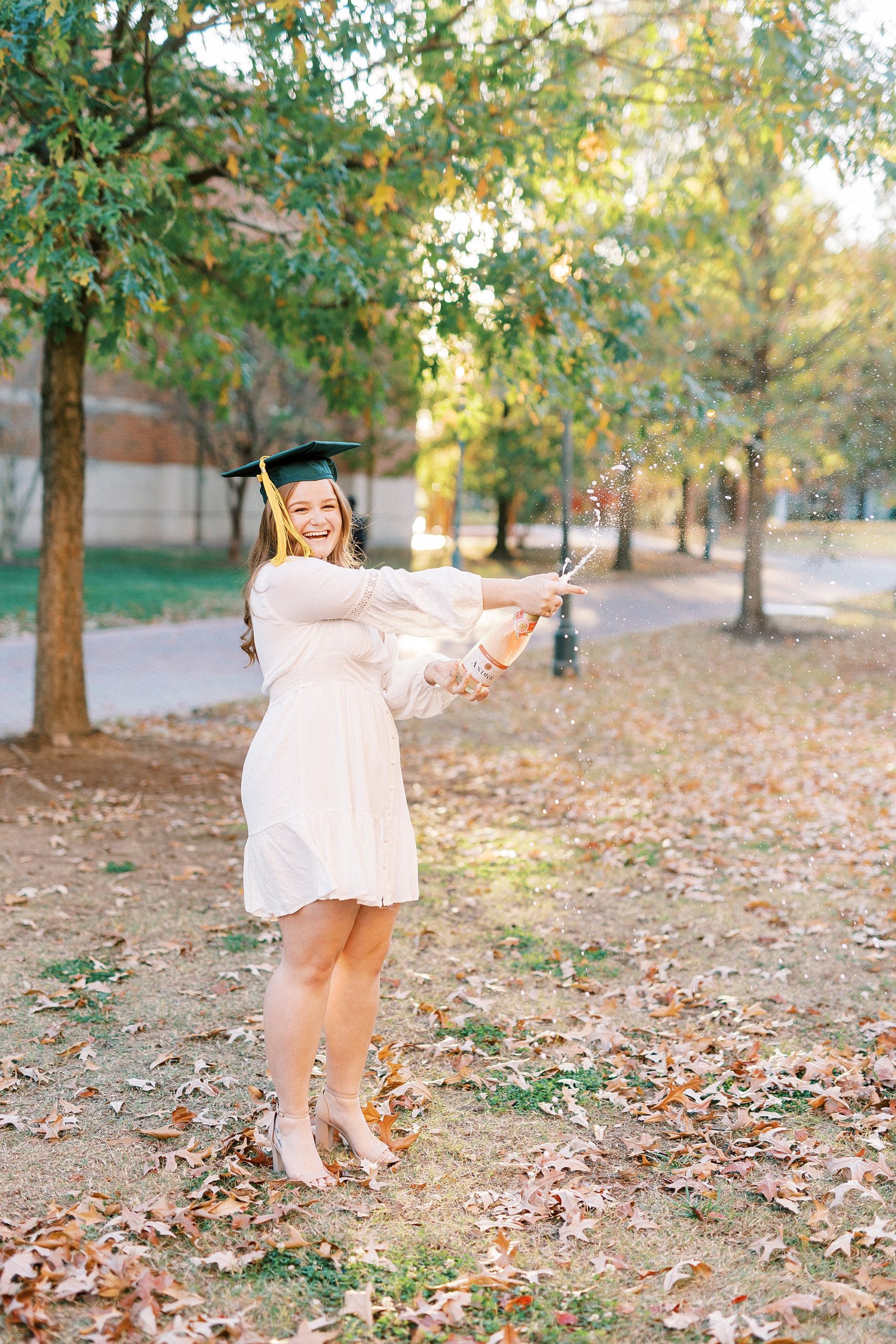 graduate in white dress with green cap pops champagne on lawn at UNC Charlotte