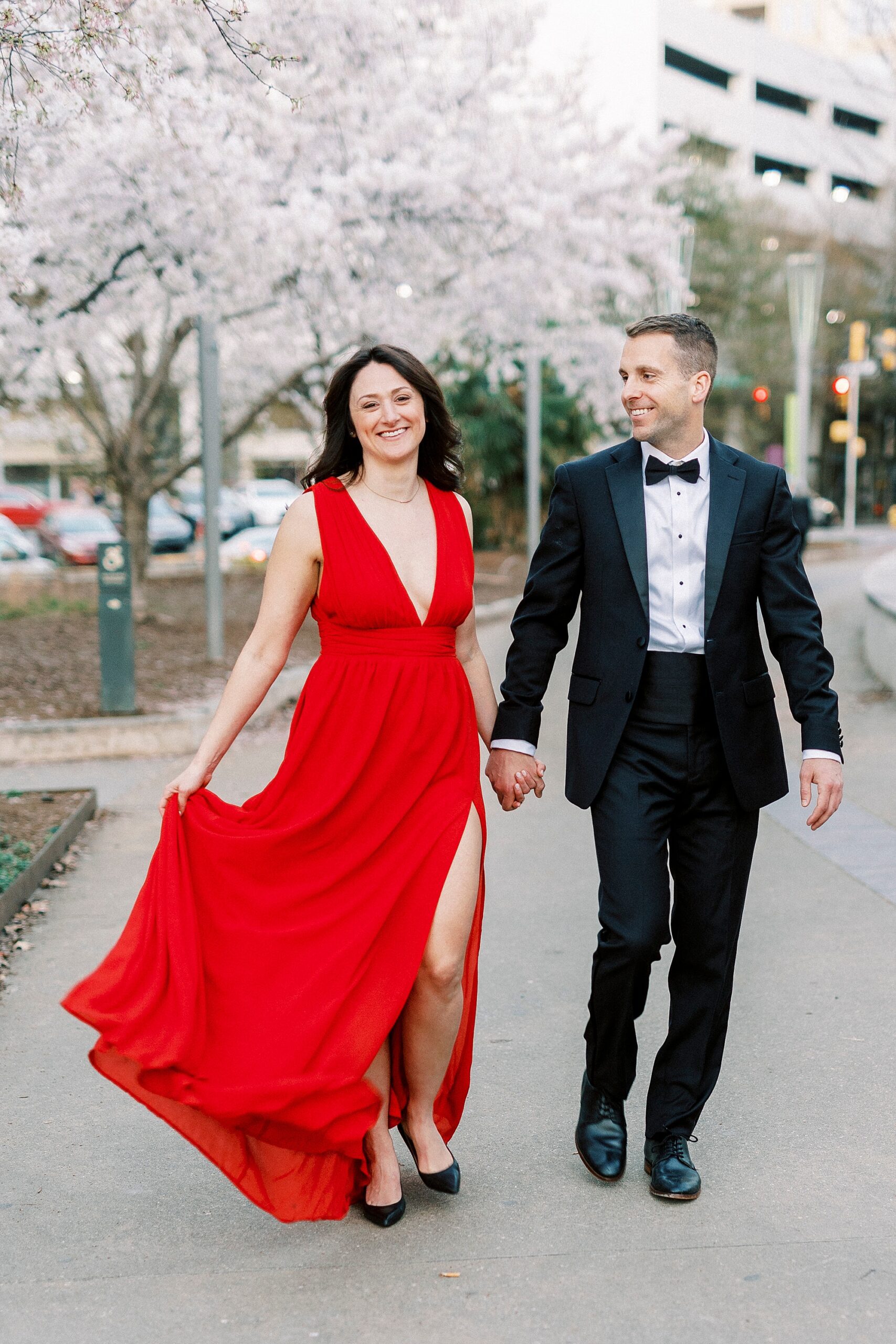 engaged couple holds hands while woman walks in red dress