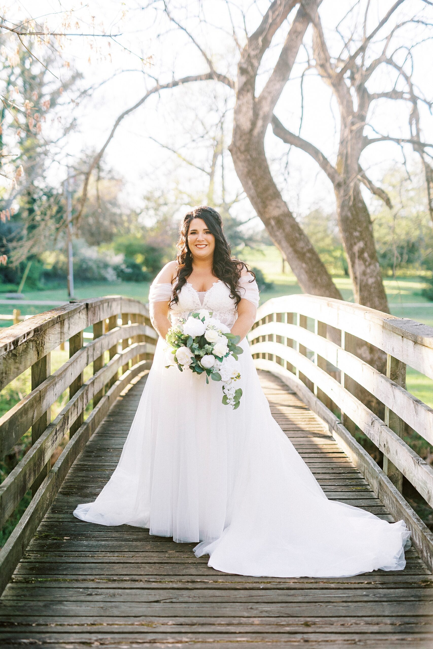 bride stands on wooden bridge holding all-white bouquet 