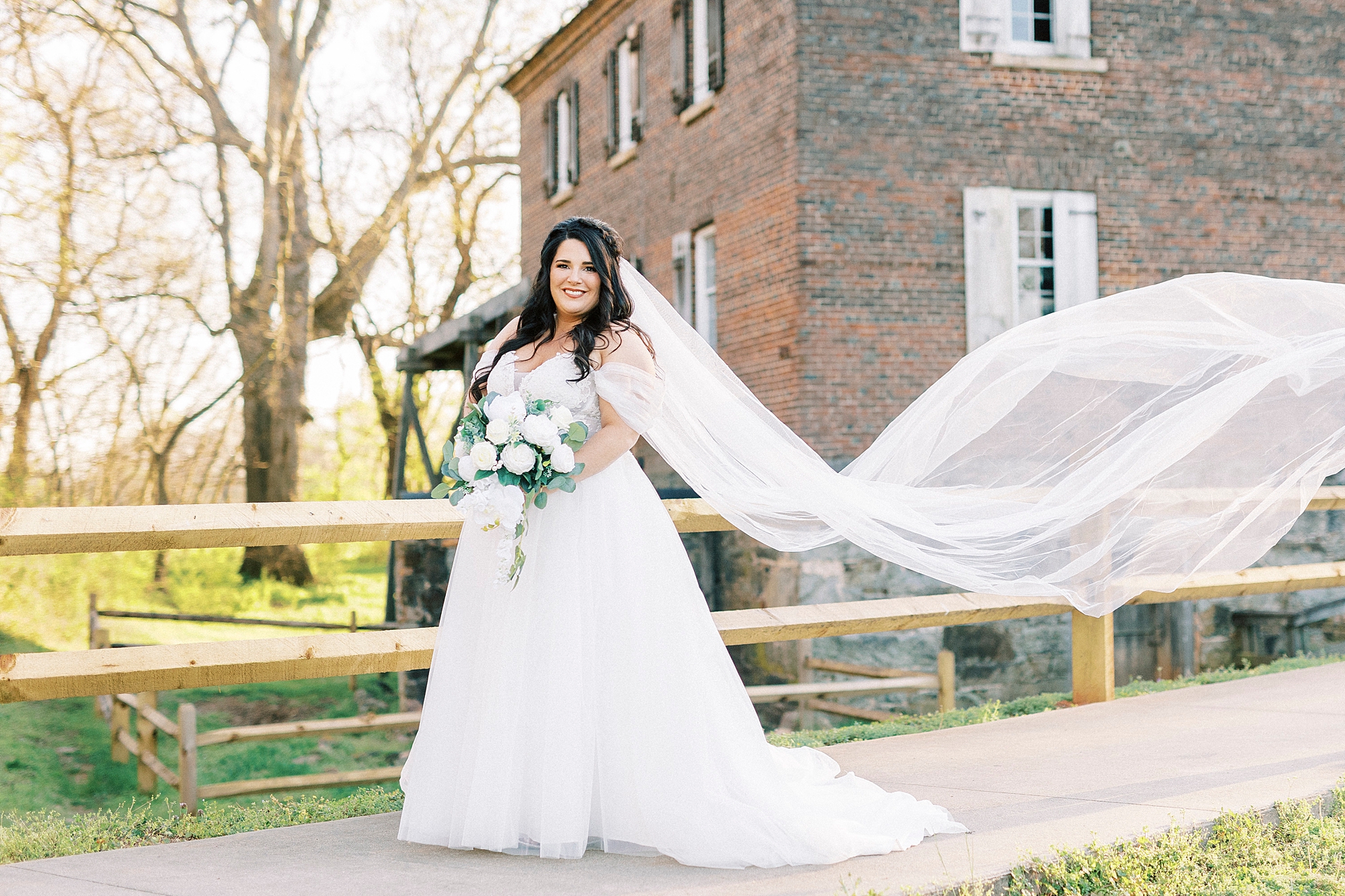 bride smiles holding bouquet of white flowers with veil floating behind her in Sloan Park