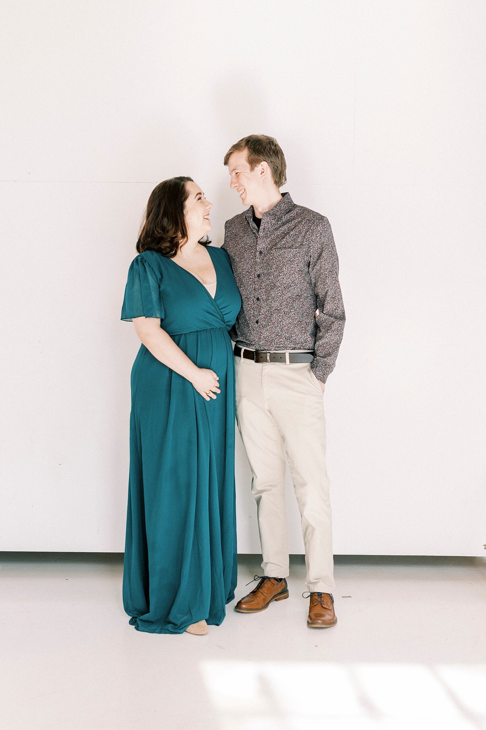 woman in teal dress holds baby bump smiling at husband during studio pregnancy announcement in Concord NC