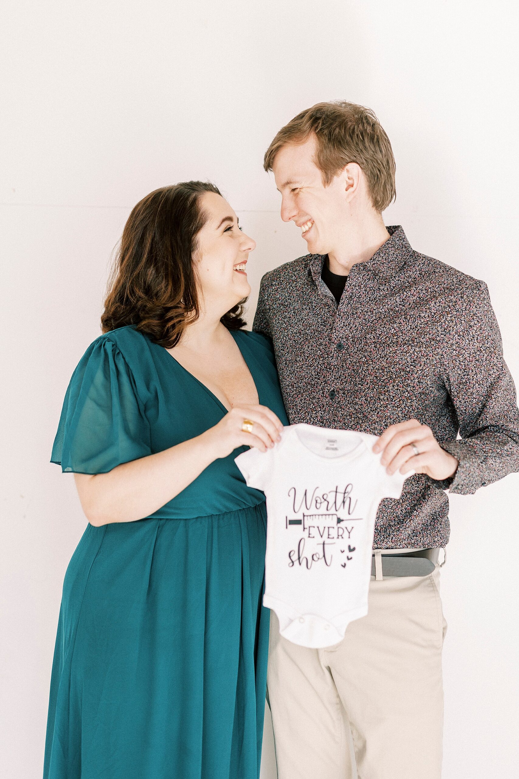 expecting parents hold baby onesie between them during studio pregnancy announcement in Concord NC