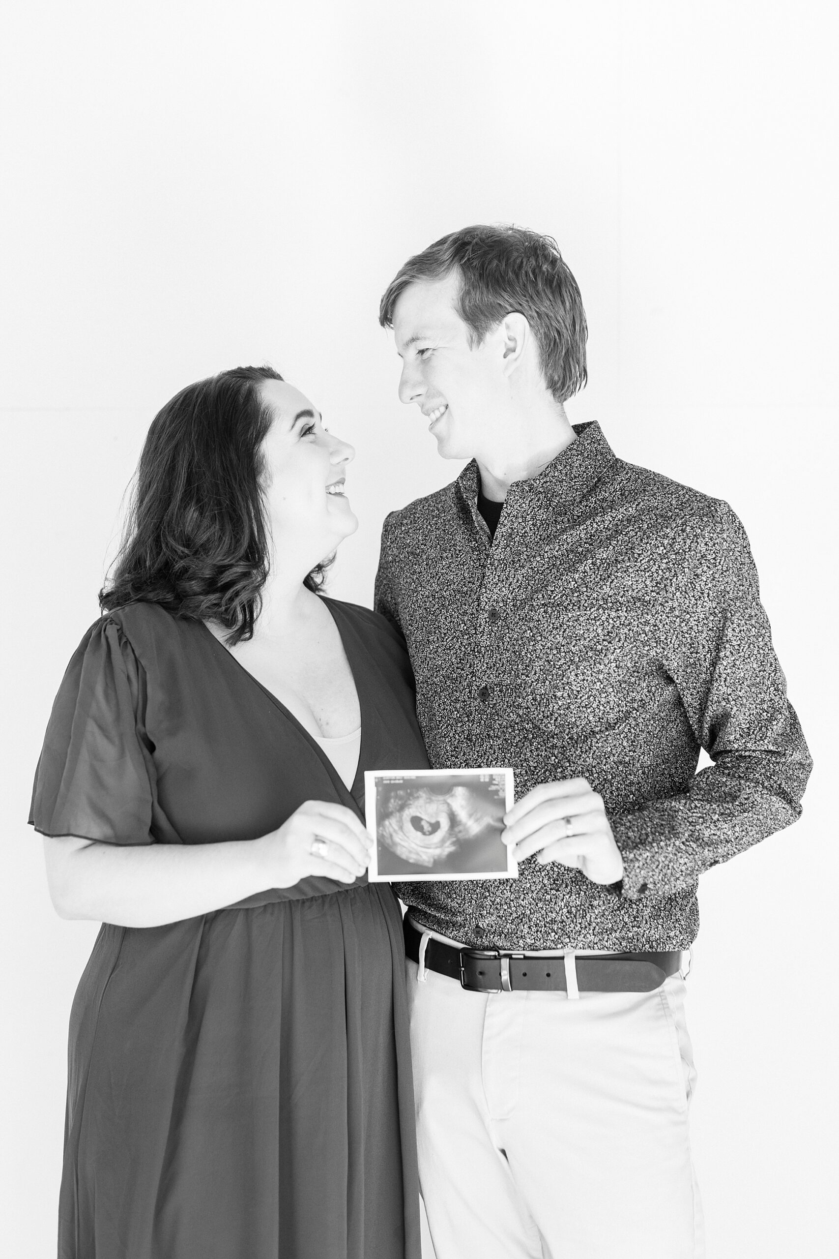 black and white portrait of couple hugging with sonogram during studio pregnancy announcement in Concord NC