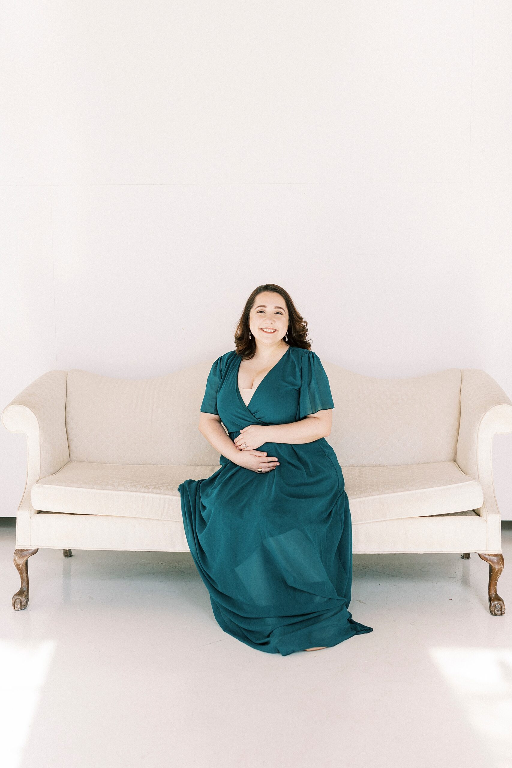 expecting mother sits in teal gown on ivory couch during studio pregnancy announcement in Concord NC