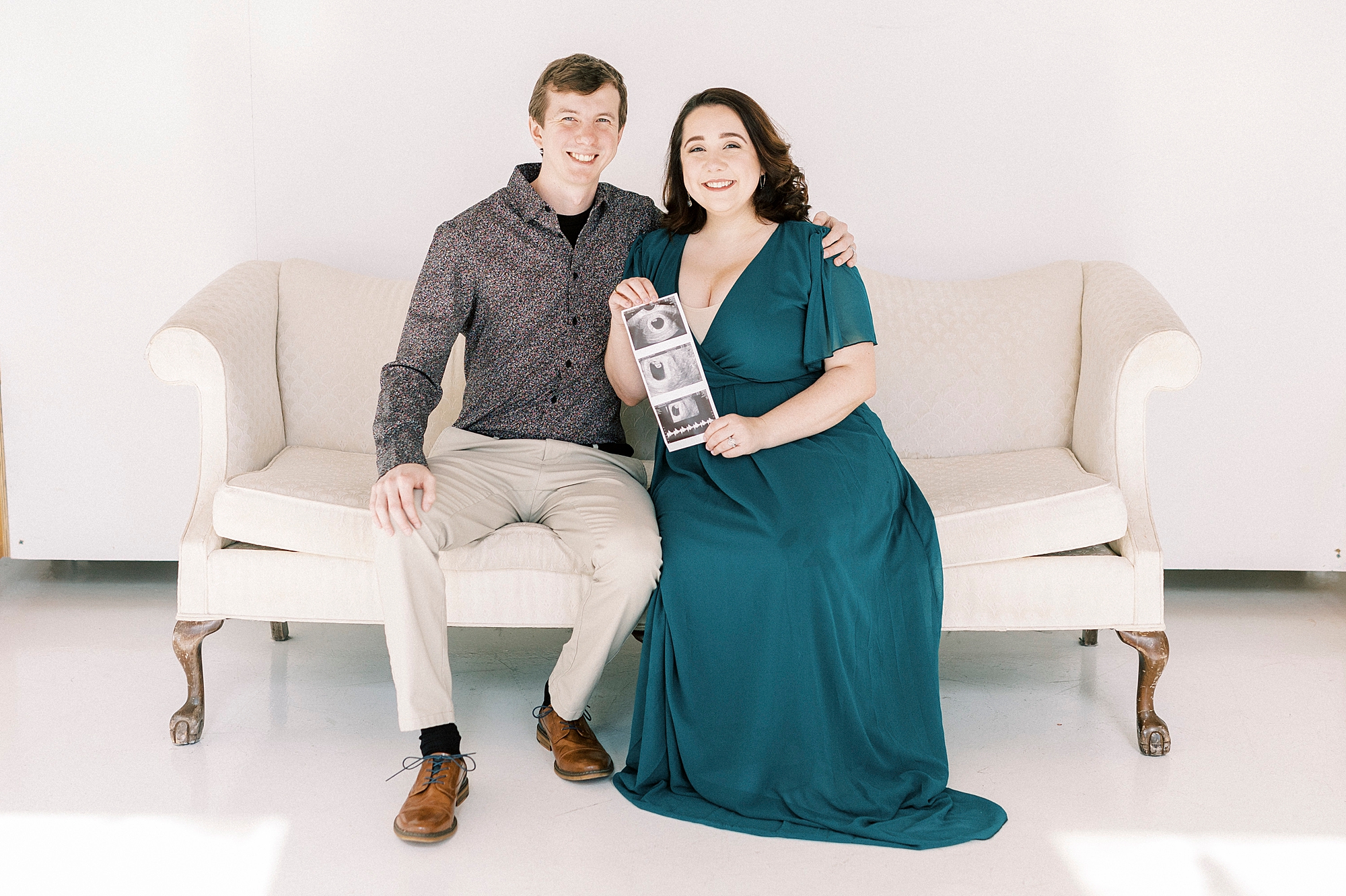 expecting parents sit on ivory couch together during studio pregnancy announcement in Concord NC