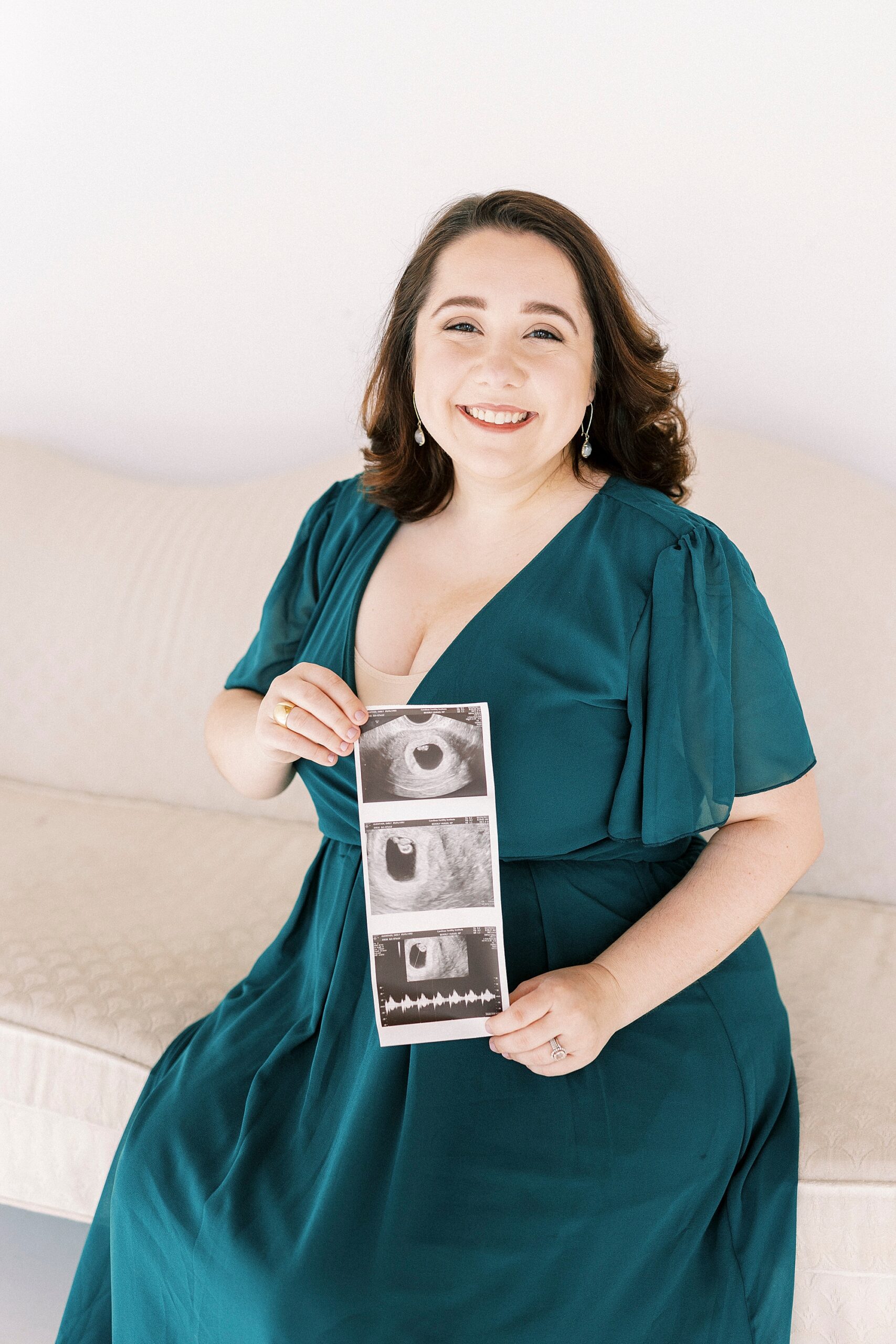 woman in teal gown holds sonogram photos on couch during studio pregnancy announcement in Concord NC