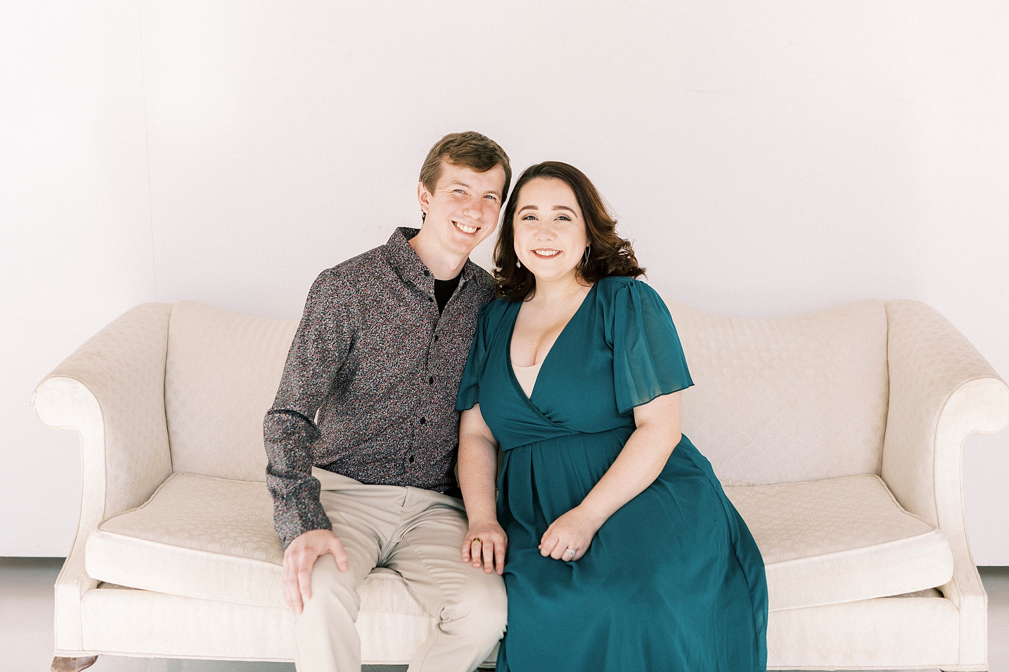 husband and wife sit on ivory couch during studio pregnancy announcement in Concord NC