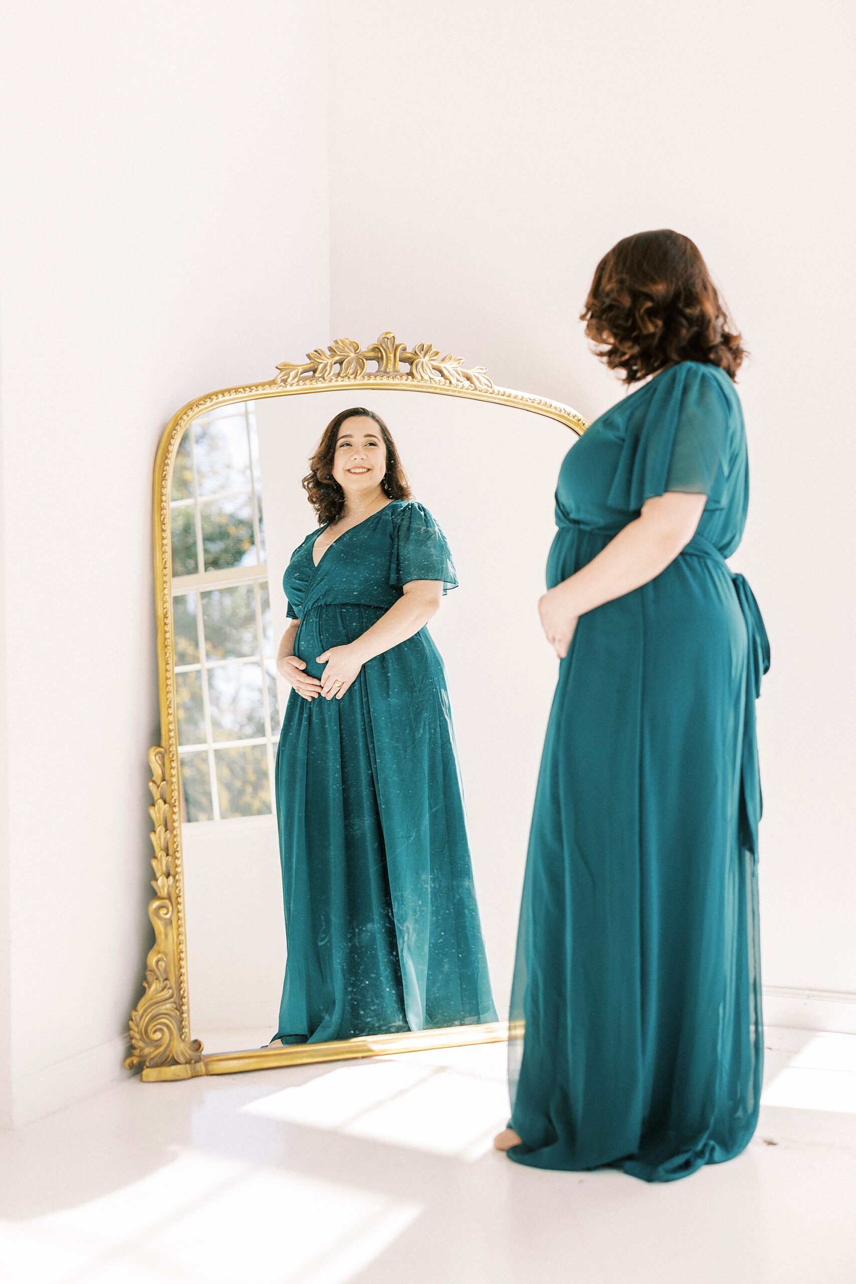 woman in teal gown holds baby bump looking in gold rimmed mirror 
