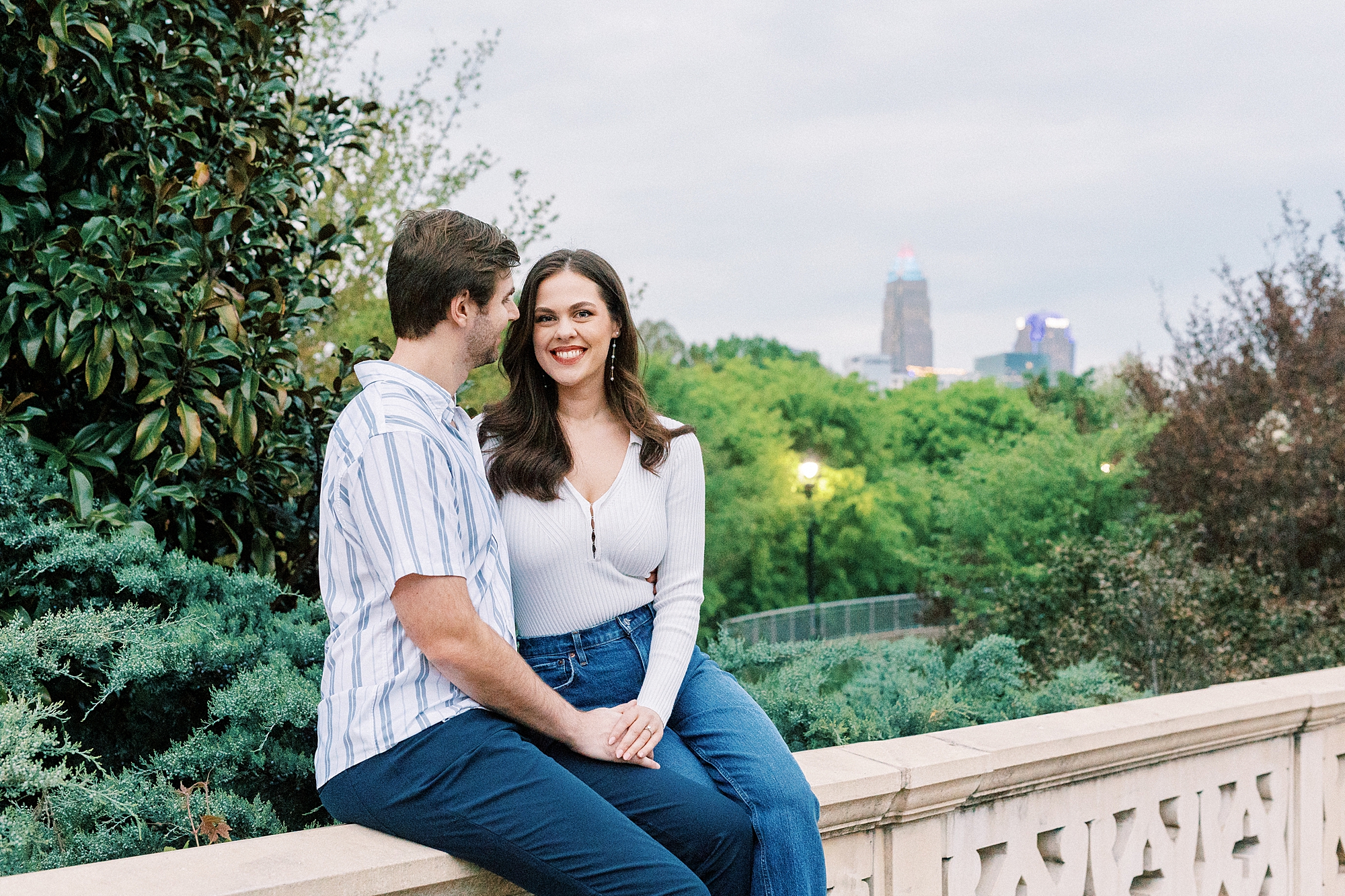 engaged couple sits on bridge with Charlotte skyline behind them 