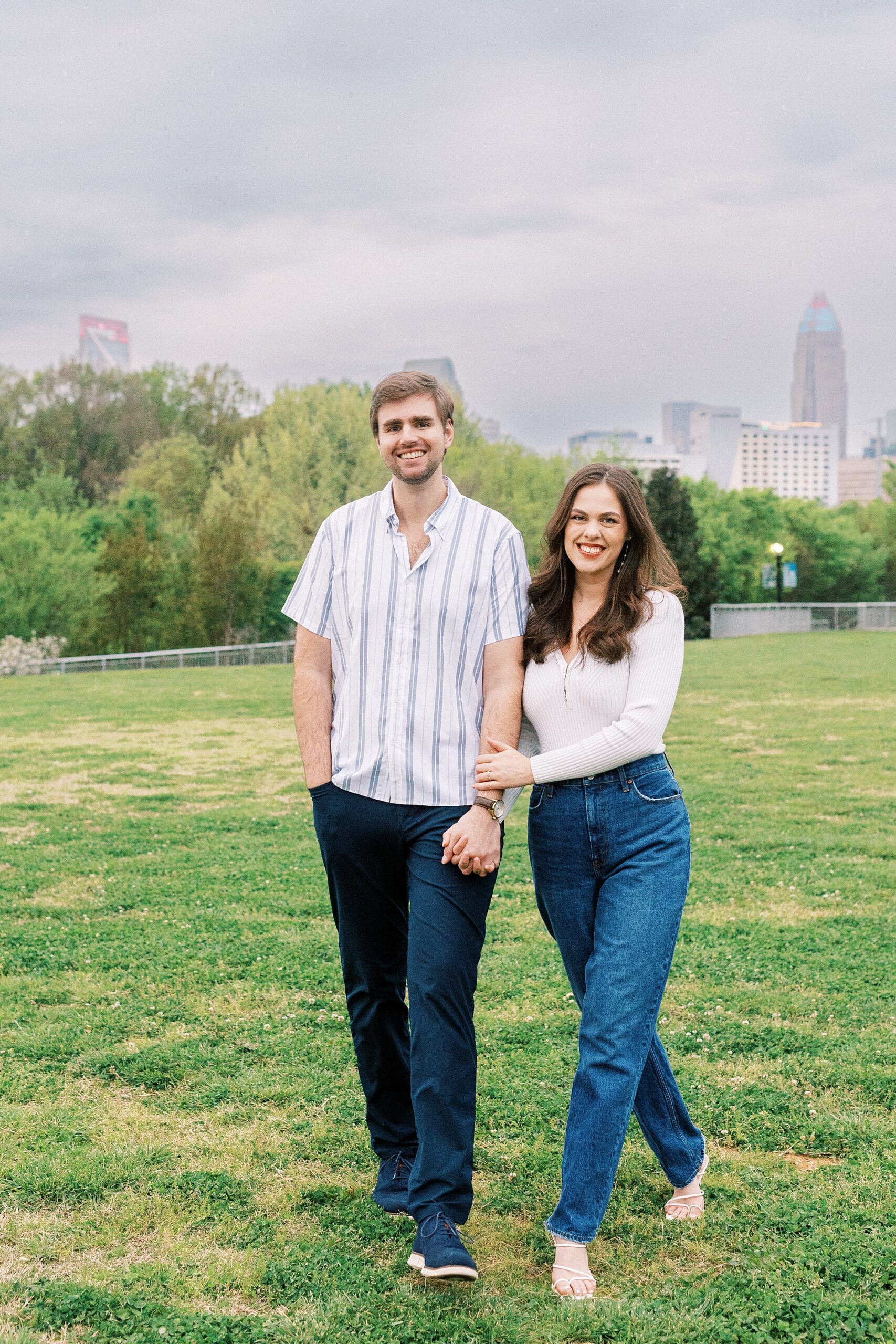engaged couple holds hands in Midtown Park with Charlotte skyline behind them 