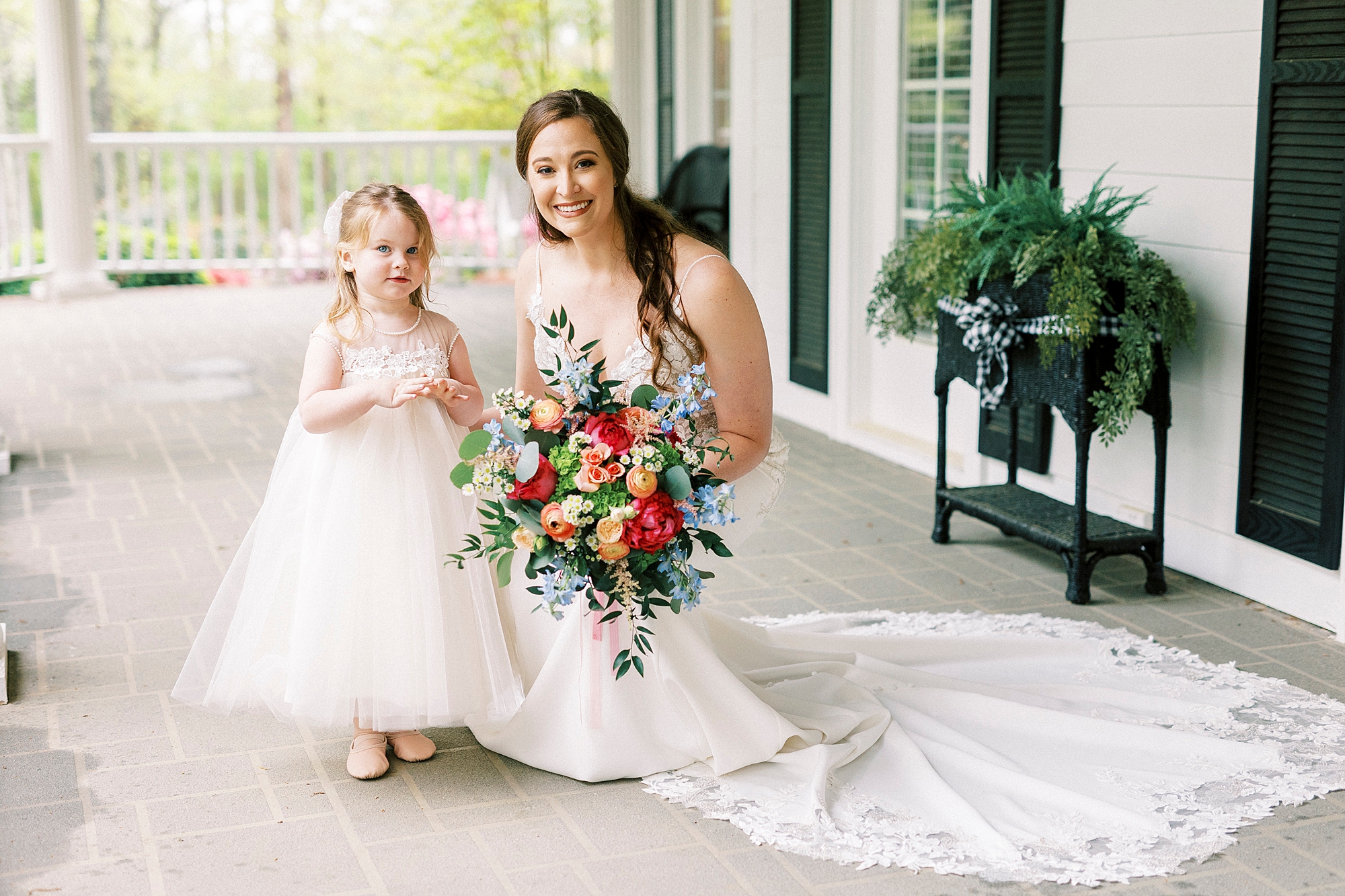 bride with bright bouquet of flowers kneels next to flower girl on porch 
